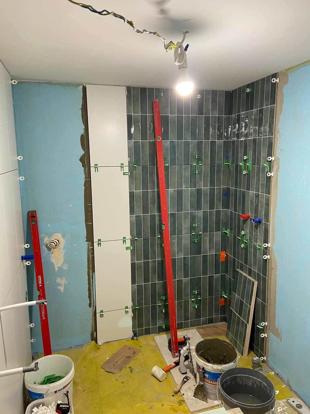 Łazienka w trakcie układania ciemnozielonych, błyszczących płytek wertykalnych, widoczne poziomice, wiadra z zaprawą, krzyżyki dystansowe i niebieska ściana w tle.