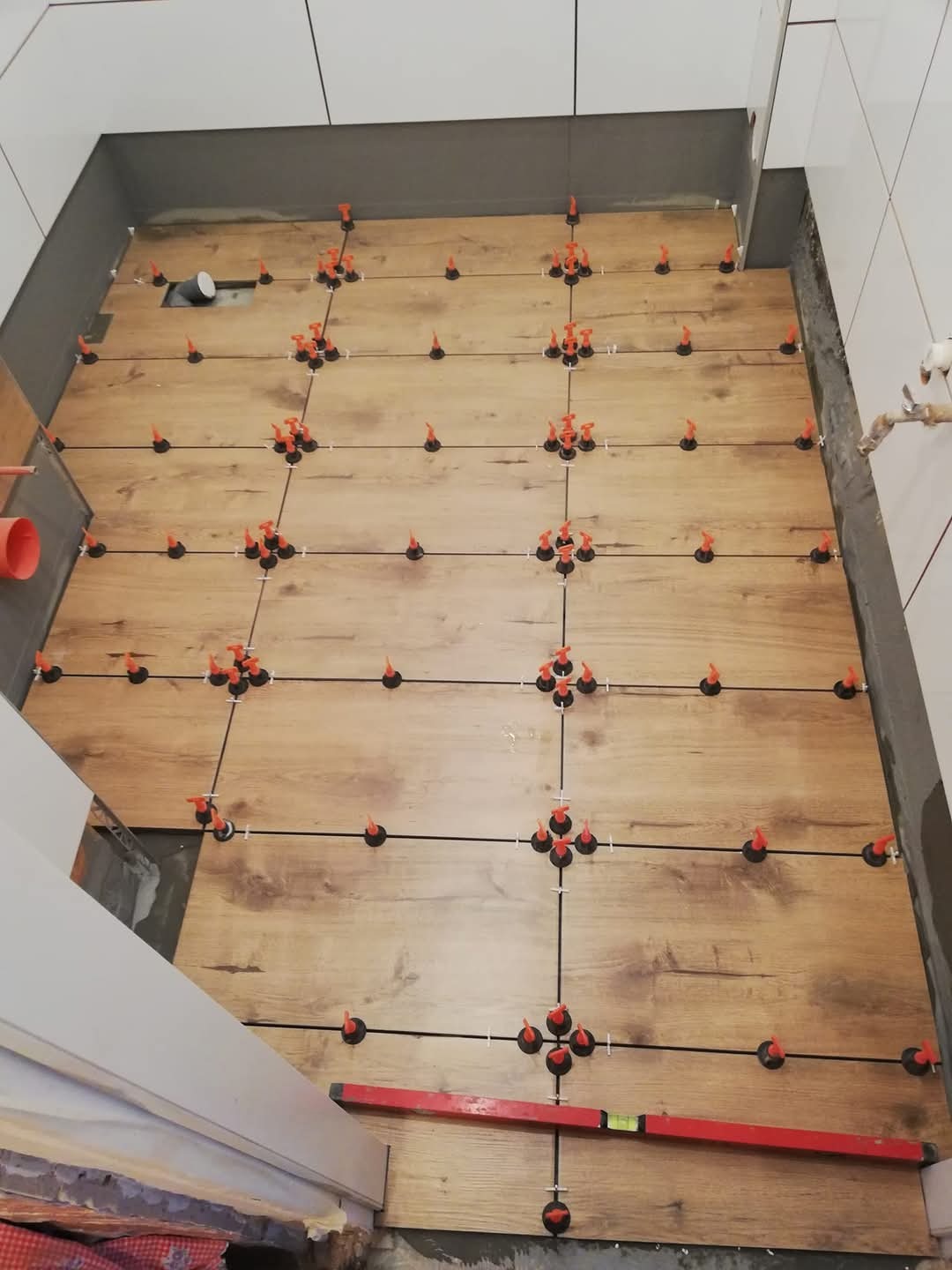 Układanie drewnopodobnych płytek podłogowych z użyciem systemu poziomowania, widoczny poziomica i fragment rury kanalizacyjnej.