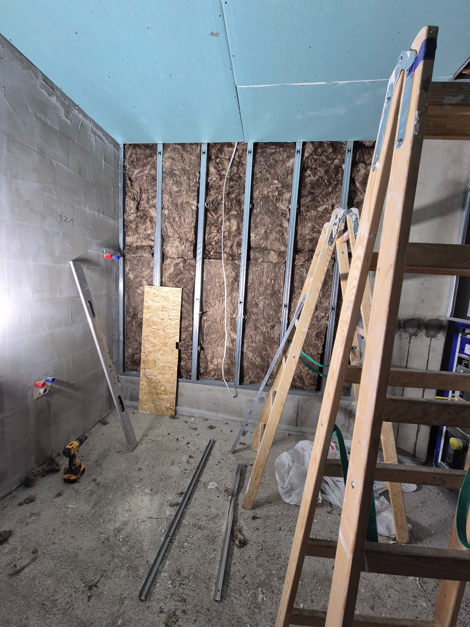 Wnętrze w trakcie prac wykończeniowych: ściana z izolacją z wełny mineralnej w metalowym stelażu, częściowo zakryta płytami gipsowo-kartonowymi, drabina drewniana, poziomica, wiertarka i profile...