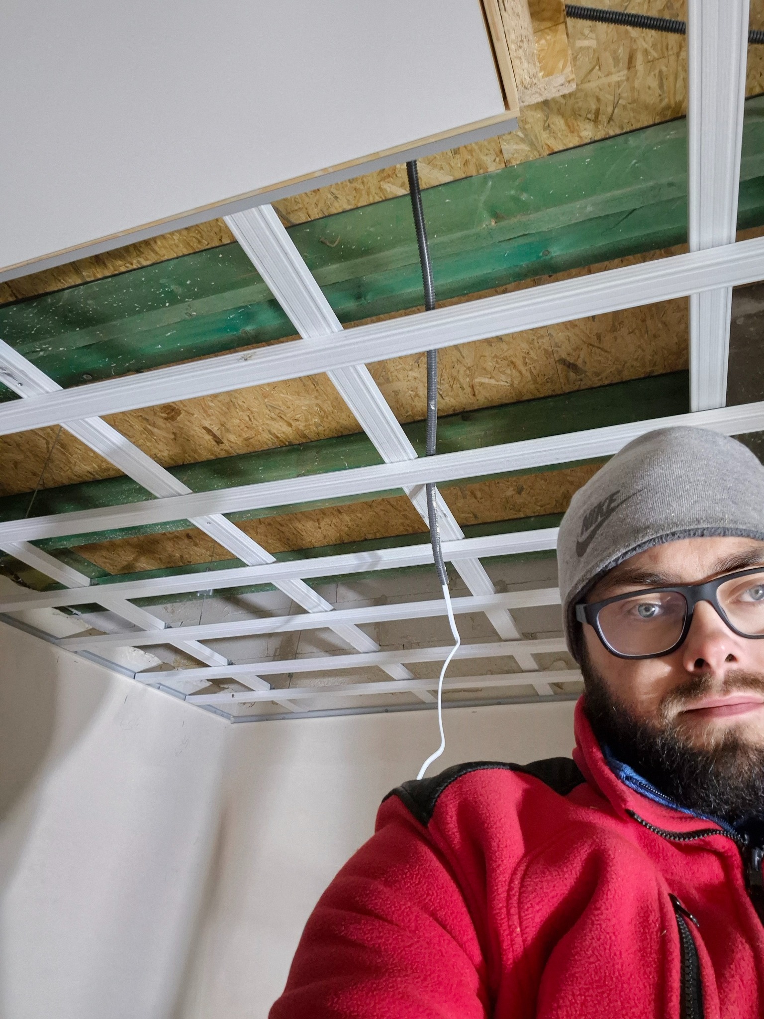 Montaż stelaża sufitu podwieszanego z widocznymi profilami, instalacją elektryczną i częściowo zamontowaną płytą gipsową, uchwycone z perspektywy wykonawcy w czerwonej polarowej kurtce i szarej...