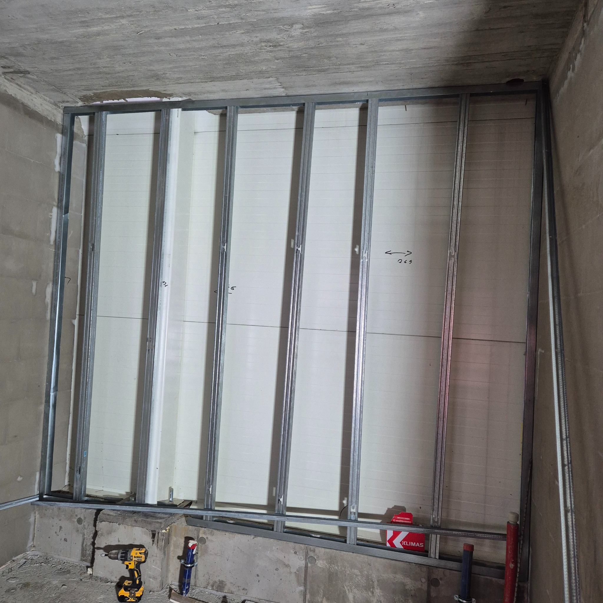 Stelaż z profili metalowych pod płytę gipsowo-kartonową przy betonowej ścianie, widoczny wkrętak akumulatorowy i opakowanie kołków montażowych.