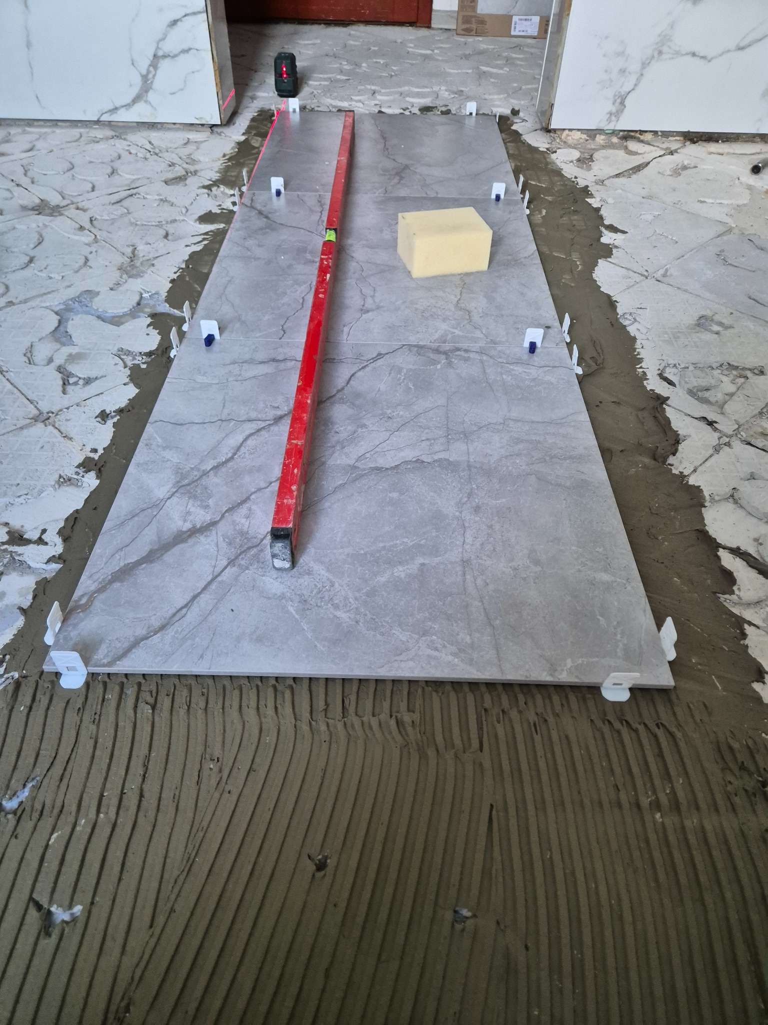 Ułożona szara płytka imitująca marmur, poziomowana czerwoną poziomicą i laserem, z użyciem białych klipsów dystansowych, na warstwie kleju z widocznymi rowkami.