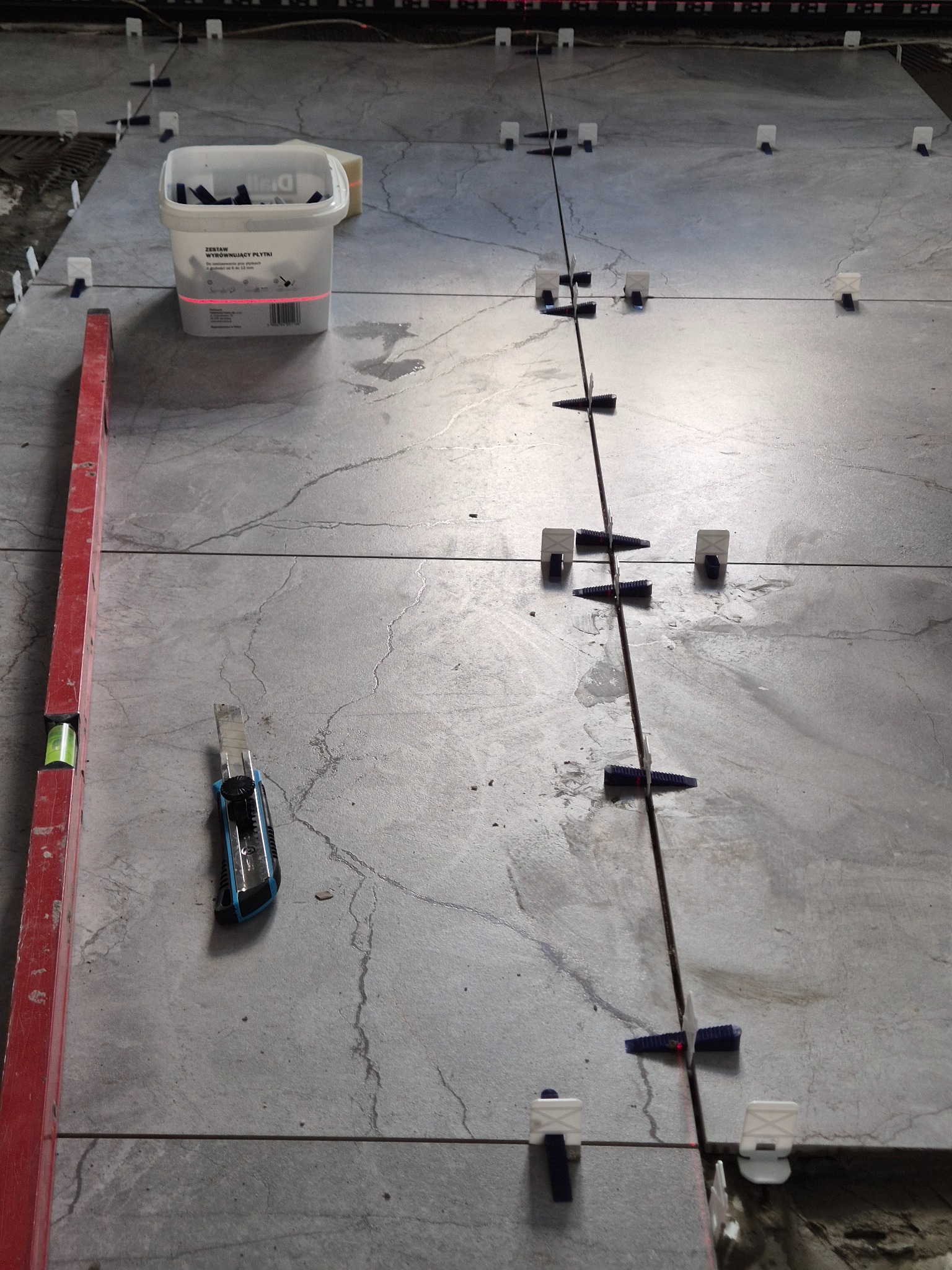 Układanie szarych płytek podłogowych z użyciem systemu poziomowania, widoczna poziomica, nożyk do cięcia płytek i wiadro z klinami.