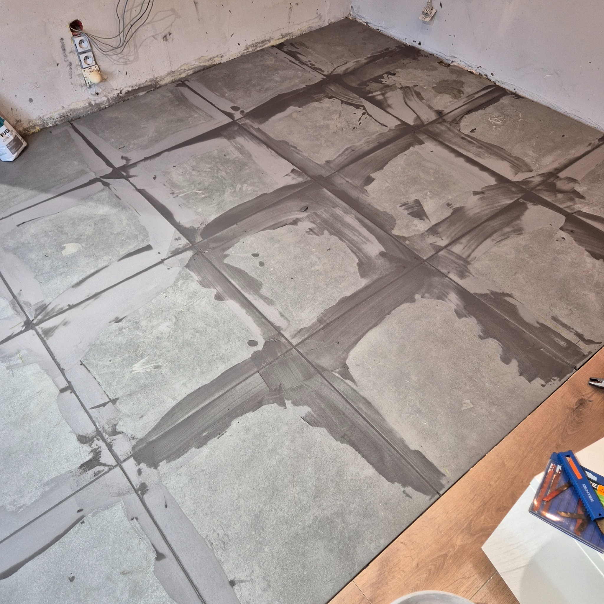 Szare płytki podłogowe w trakcie fugowania, widoczne świeże, ciemne fugi, przy ścianie puszka z fugą, w tle surowa ściana z wystającymi kablami.