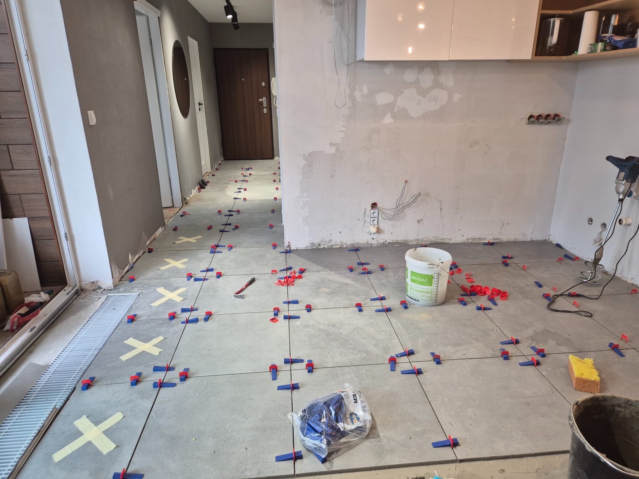 Układanie szarych płytek podłogowych z użyciem systemu poziomowania płytek (klipsy i kliny) w trakcie prac wykończeniowych w mieszkaniu. Widoczne narzędzia i wiadro z klejem.