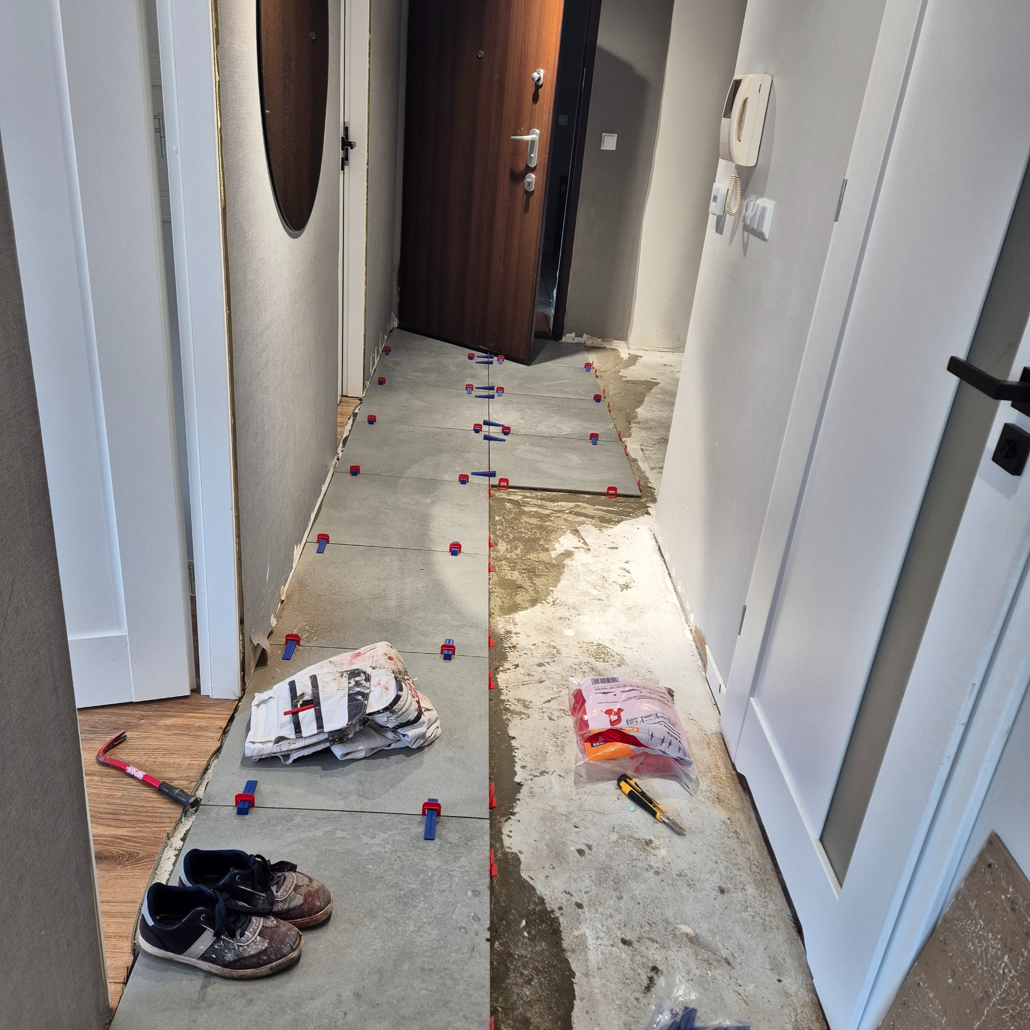 Układanie szarych płytek podłogowych w korytarzu z użyciem klipsów poziomujących, widoczne narzędzia, paczka z klipsami, oraz para roboczych butów.