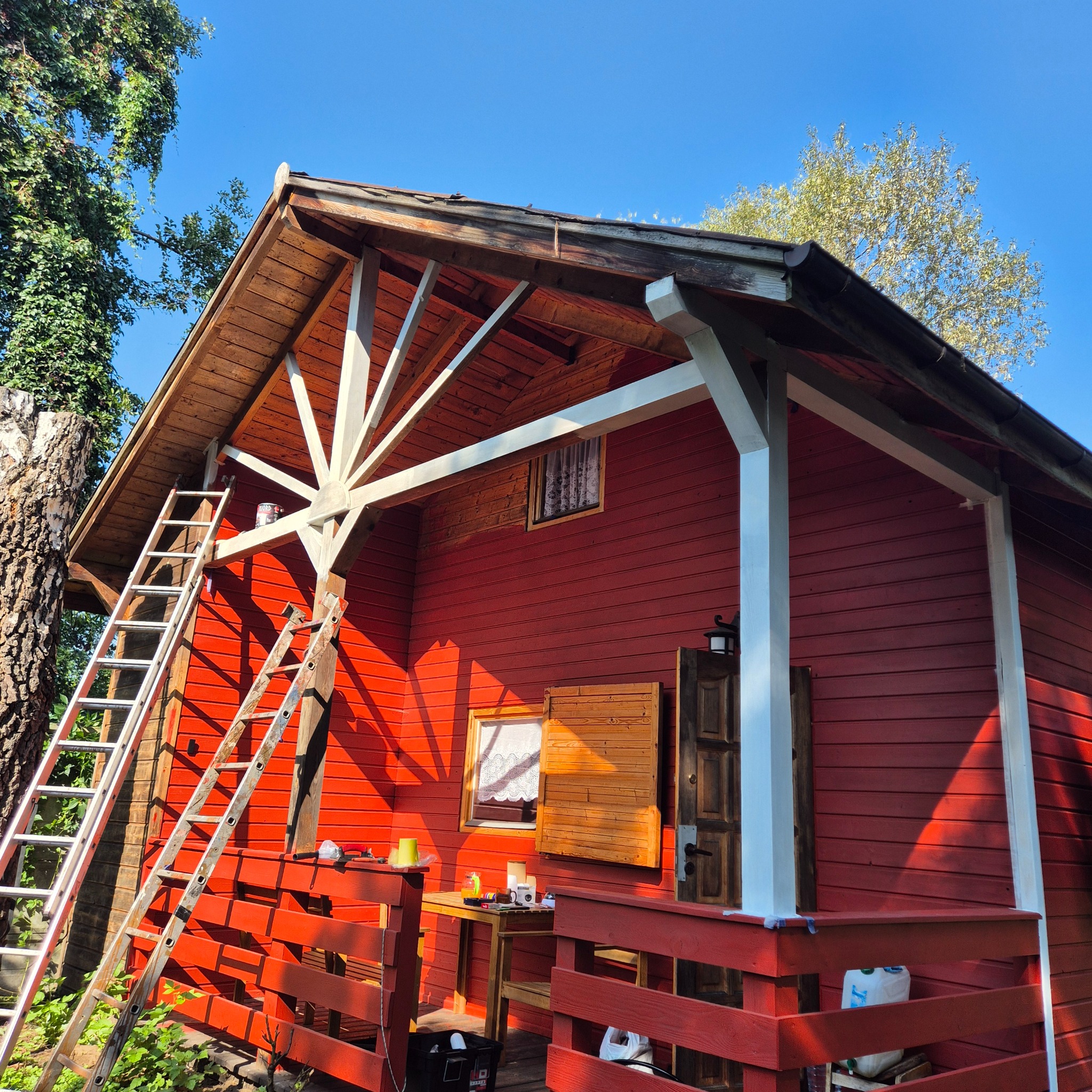 Czerwony drewniany domek w trakcie malowania, z drabiny opartej o ścianę, częściowo pomalowane elementy konstrukcyjne na biało, widoczne narzędzia i puszki z farbą.