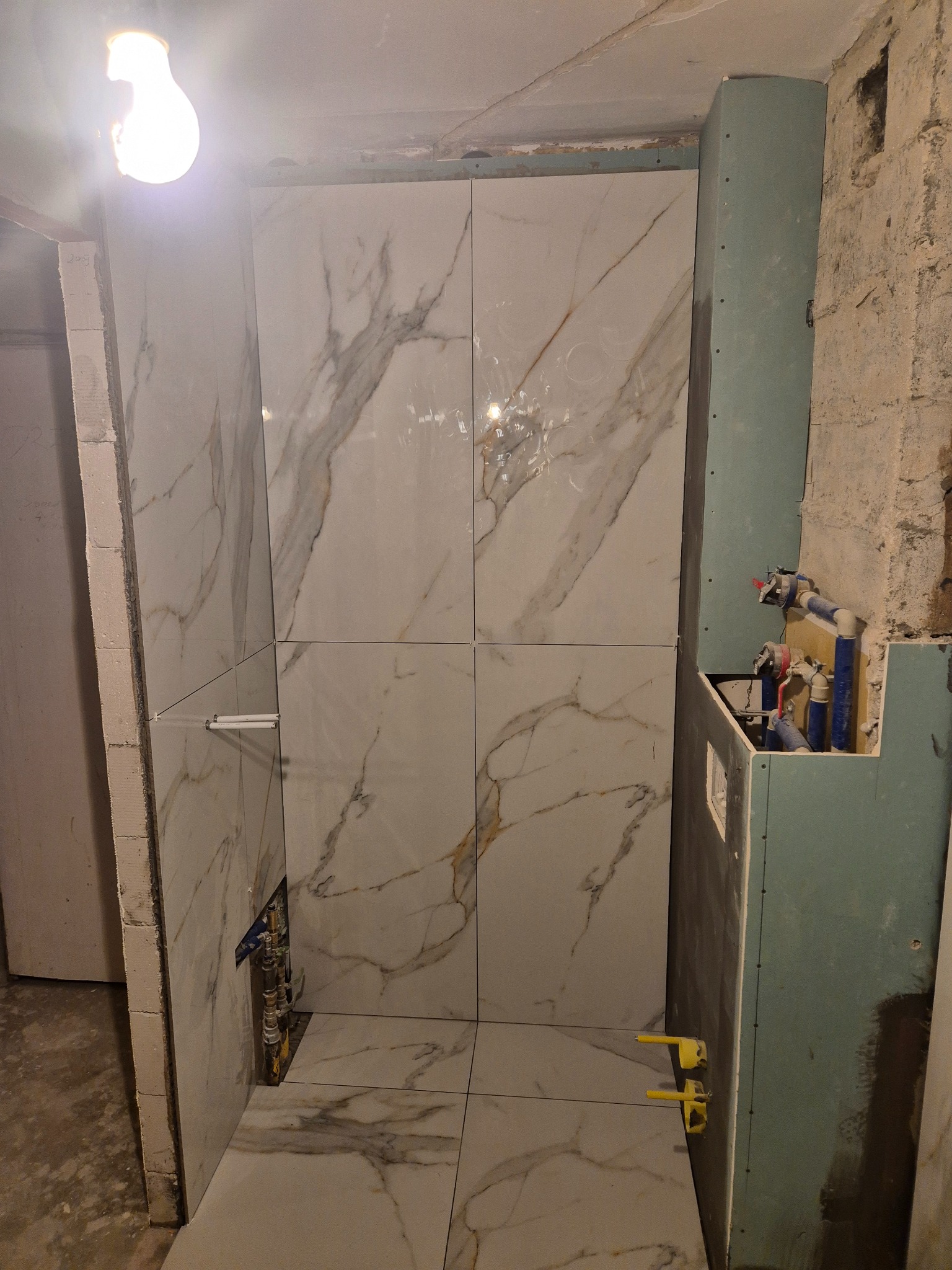 Fragment łazienki w trakcie układania płytek imitujących marmur, widoczne krzyżyki dystansowe, instalacja wodna w ścianie oraz surowy stan ścian.
