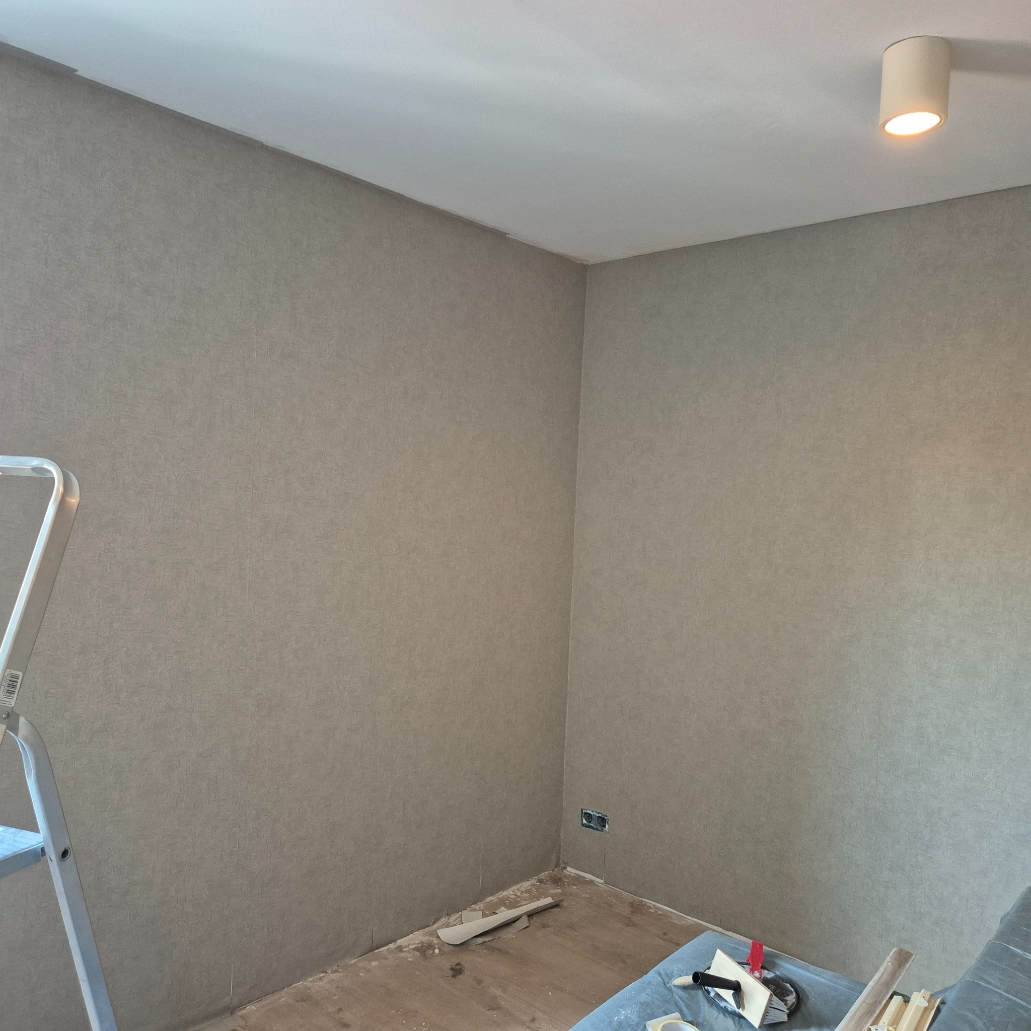 Ściany w pokoju po nałożeniu gładzi, widoczny fragment drabiny malarskiej, podłoga częściowo zabezpieczona folią, na folii narzędzia do nakładania gładzi.