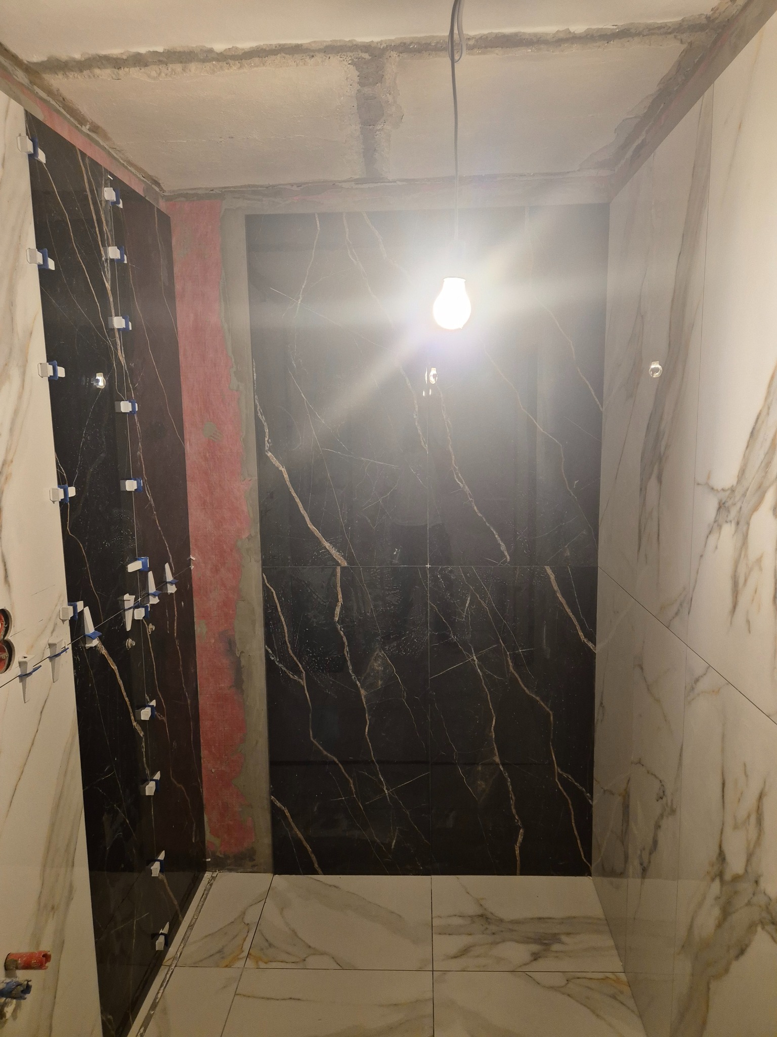 Łazienka w trakcie układania płytek imitujących marmur, widoczne białe płytki z szarymi żyłami i czarne płytki ze złotymi żyłami, krzyżyki dystansowe na ścianie, surowy sufit i wisząca żarówka.