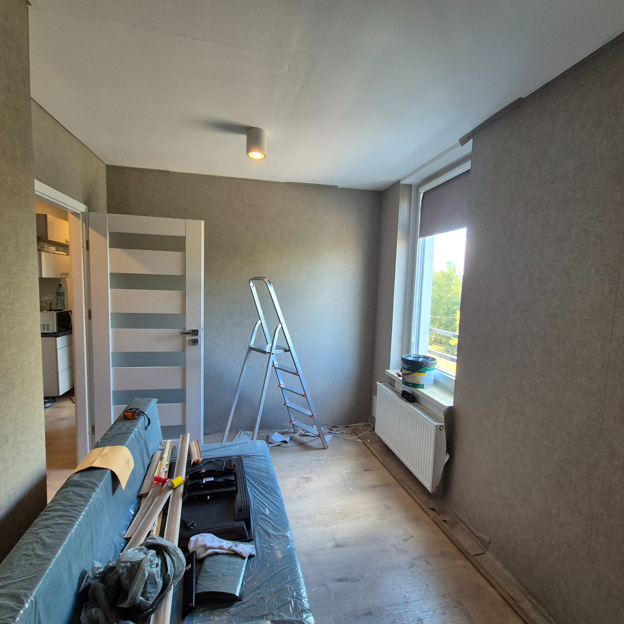 Pokój w trakcie odświeżania: drabina oparta o ścianę z widocznymi puszkami farby na parapecie, zabezpieczona folią kanapa z listwami przypodłogowymi, otwarte drzwi do kuchni w tle.