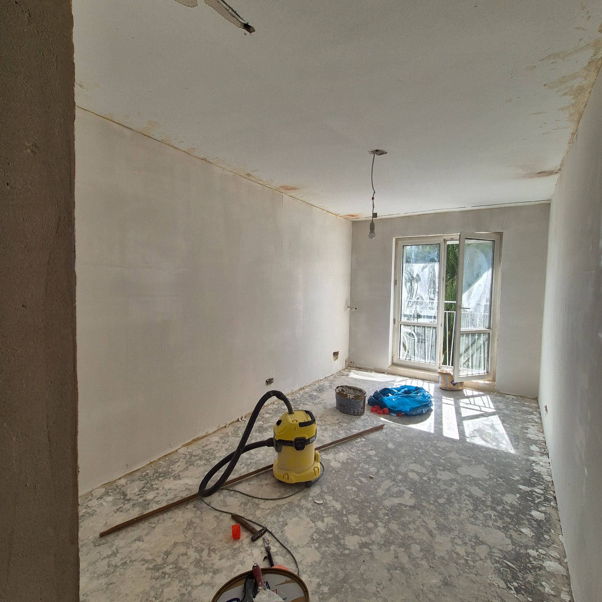 Pokój w trakcie prac przygotowawczych: ściany po nałożeniu warstwy wyrównującej, podłoga zabezpieczona przed malowaniem, widoczny odkurzacz przemysłowy, narzędzia i wiadro z pozostałościami...