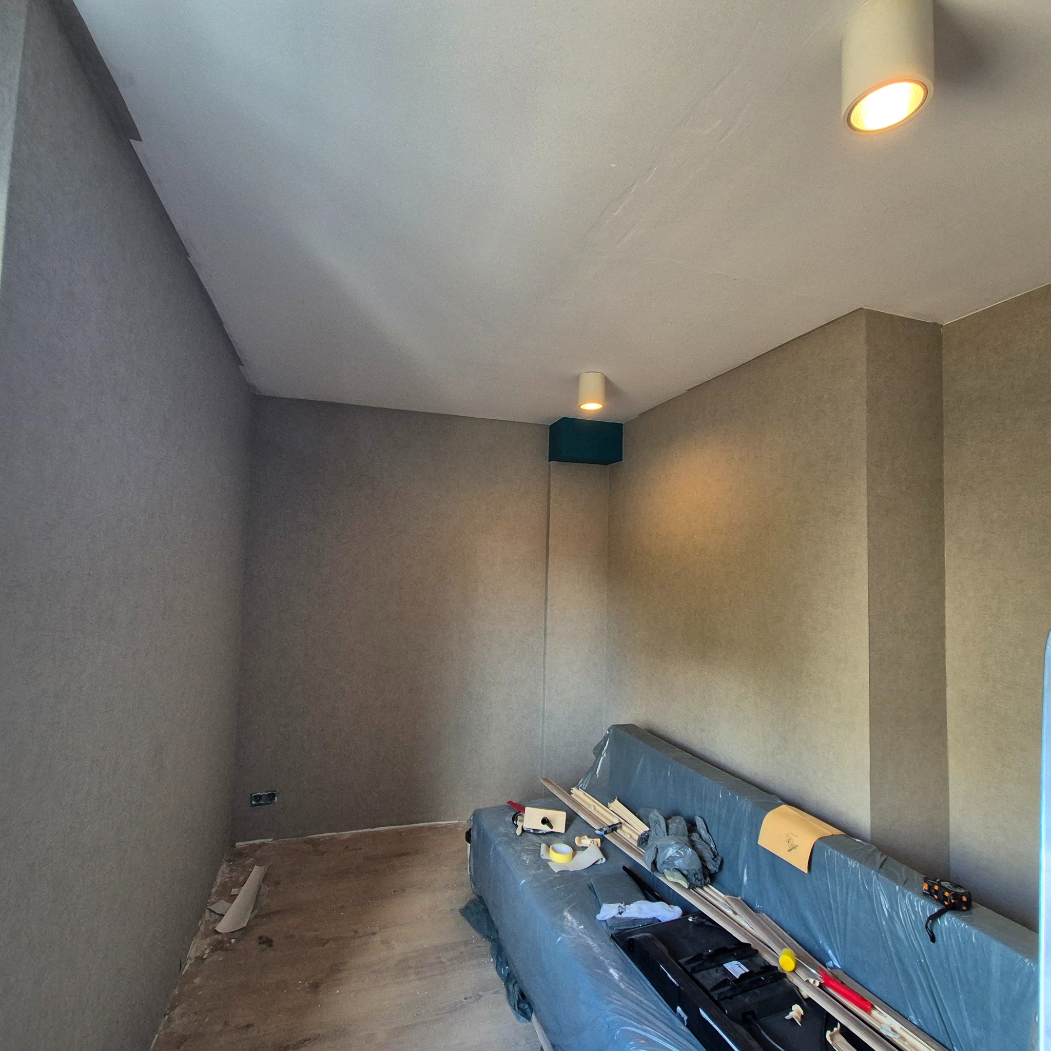 Pokój w trakcie prac wykończeniowych, ściany i sufit po wstępnym wyrównaniu, widoczne niedoskonałości i ślady po szpachlowaniu, podłoga częściowo zakryta, narzędzia i materiały budowlane...