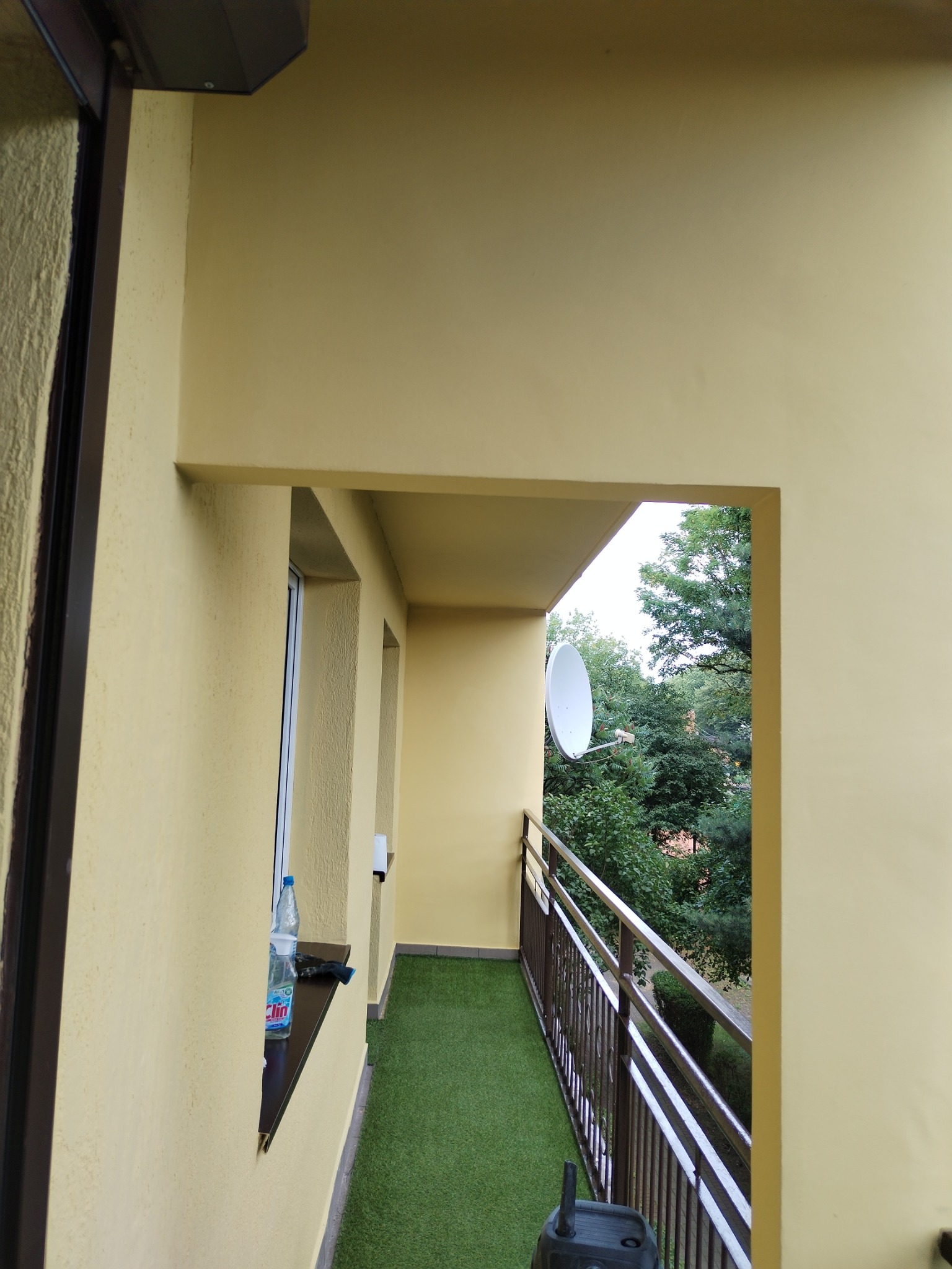 Żółty balkon z sztuczną trawą, widok na zieleń drzew i antenę satelitarną, butelka wody na parapecie.