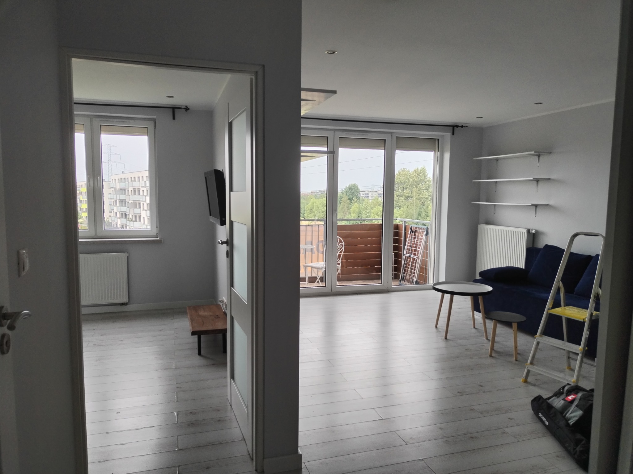 Wykończone wnętrze mieszkania w Mikołowie po remoncie, widok z perspektywy drzwi do drugiego pokoju, szare ściany, jasna podłoga, okna balkonowe, niebieska sofa i drabina malarska.