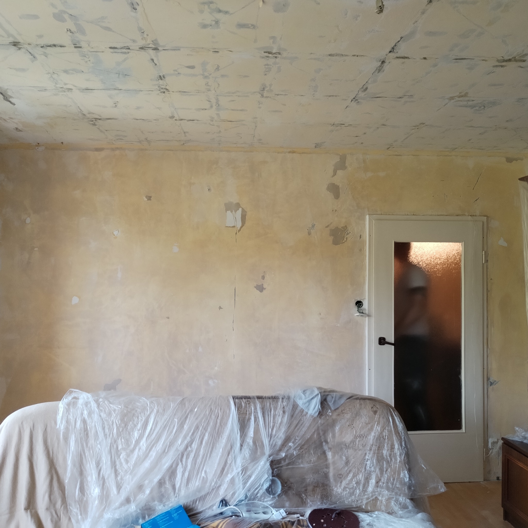 Pokój w trakcie przygotowań do malowania: surowa ściana z widocznymi śladami po szpachlowaniu, sufit z niedokończoną gładzią, meble zabezpieczone folią malarską, w tle widoczne zarys sylwetki osoby...