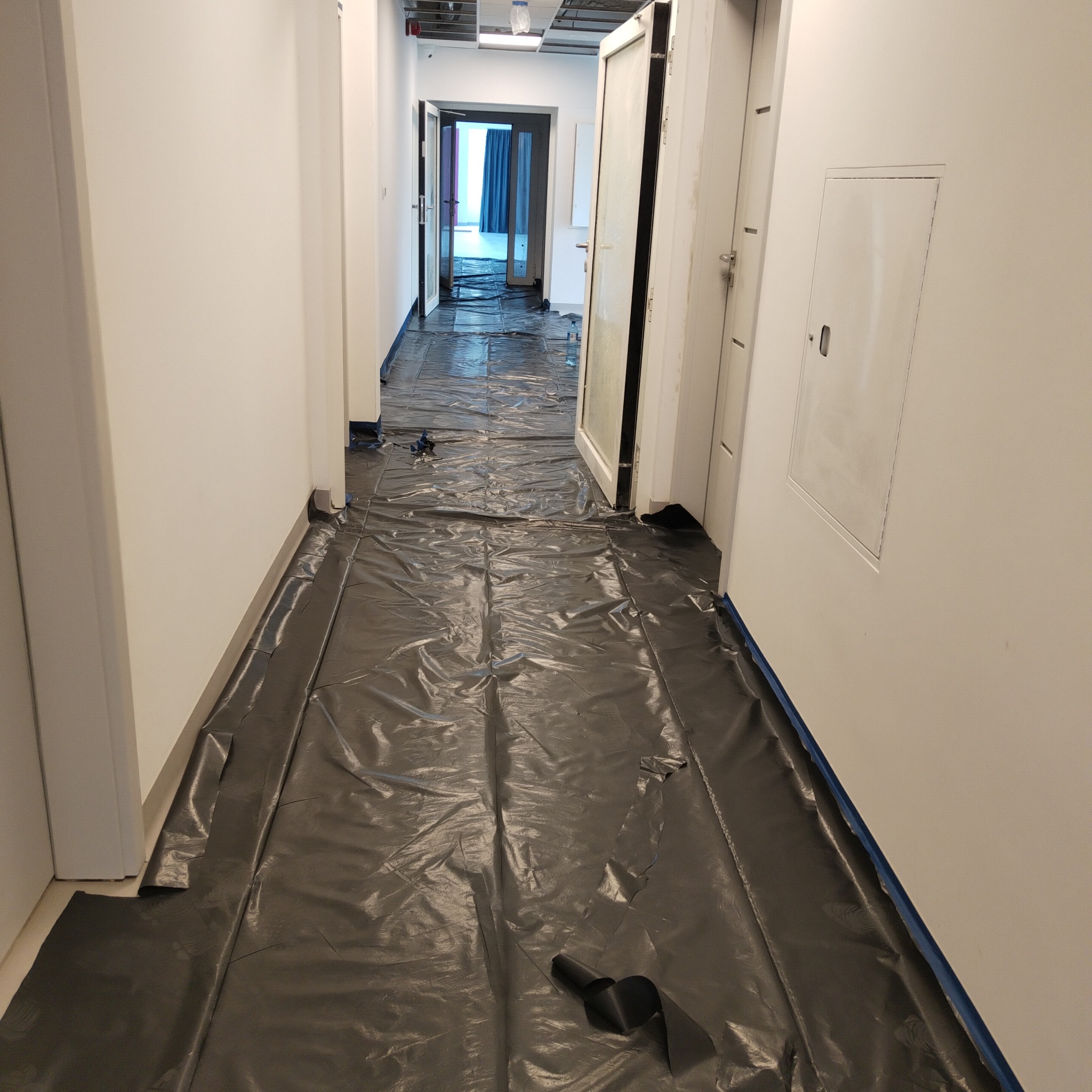 Korytarz zabezpieczony folią malarską i taśmą malarską przed rozpoczęciem prac wykończeniowych. Widoczne białe ściany i sufit podwieszany.