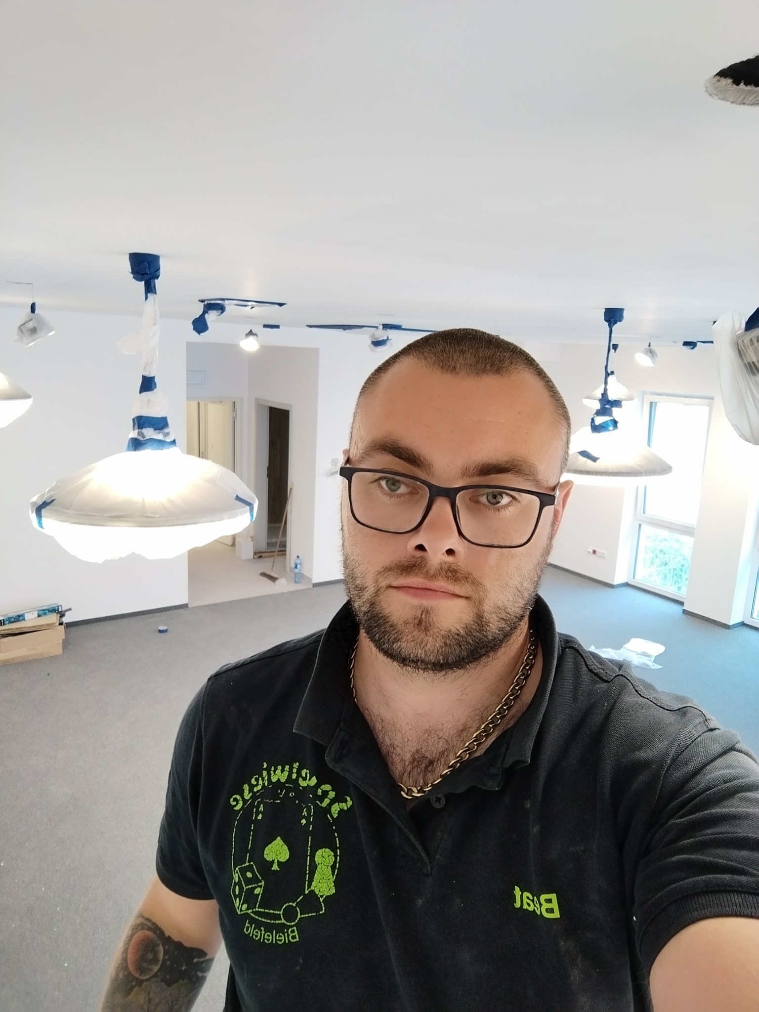 Mężczyzna w okularach, ubrany w czarną koszulkę polo z logo, robi selfie na tle pomieszczenia w trakcie prac przygotowawczych do malowania: lampy zabezpieczone folią, widoczne ślady po...