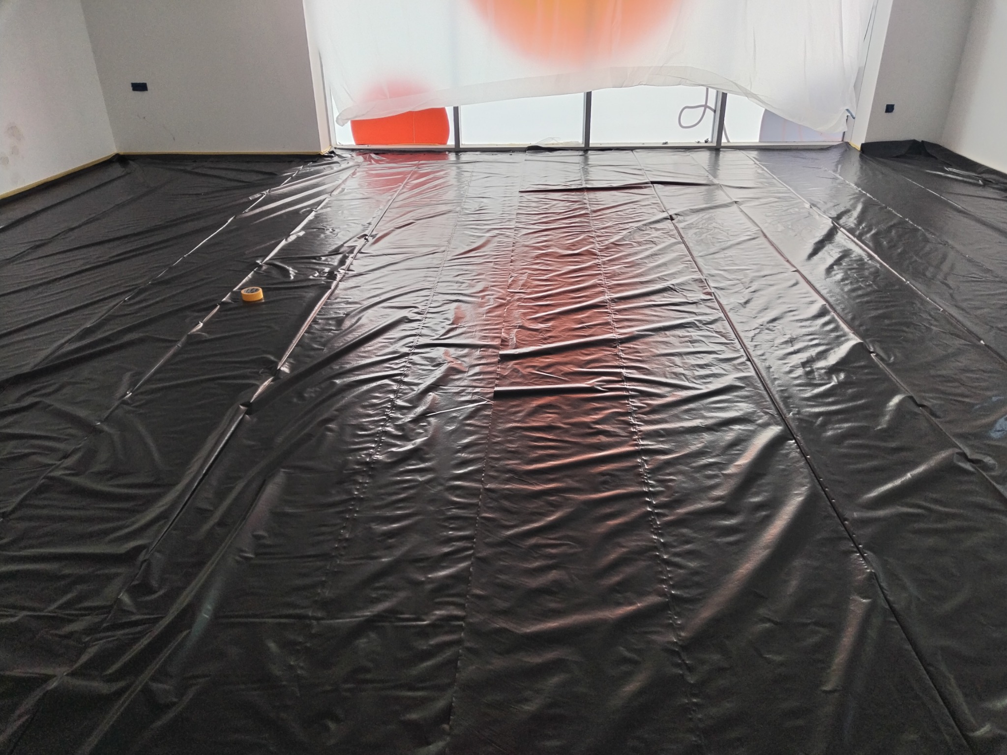 Podłoga zabezpieczona czarną folią malarską, przy krawędzi ściany taśma malarska, widoczna rolka taśmy na folii, okno zasłonięte folią ochronną.