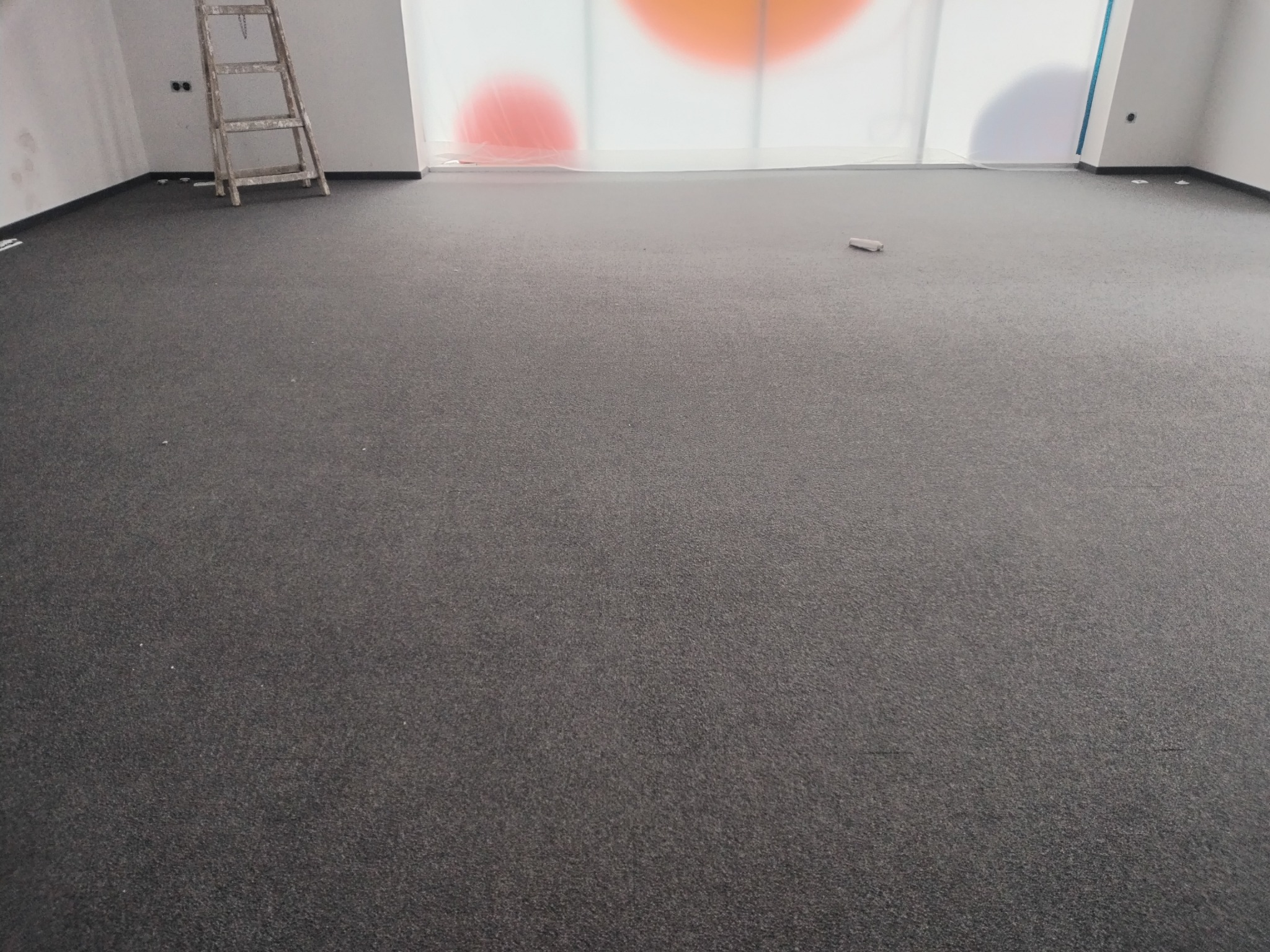 Wykończone wnętrze z nową, ciemnoszarą wykładziną dywanową, drabina oparta o ścianę, okno zasłonięte folią ochronną z kolorowymi okręgami.