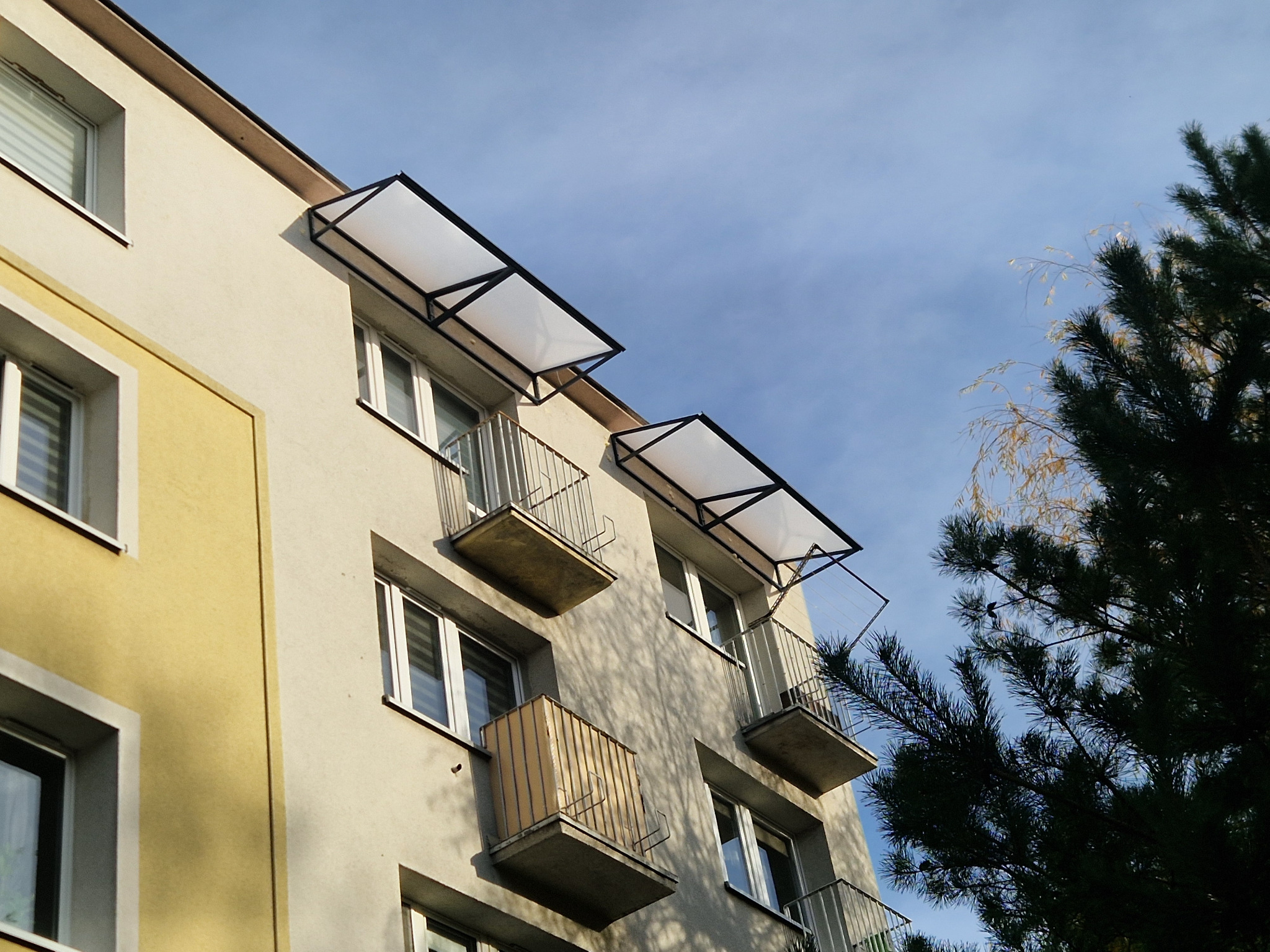 Balkony z metalowymi balustradami i zadaszeniami z przezroczystego tworzywa, zamontowane na elewacji bloku mieszkalnego, widziane z dołu pod kątem.