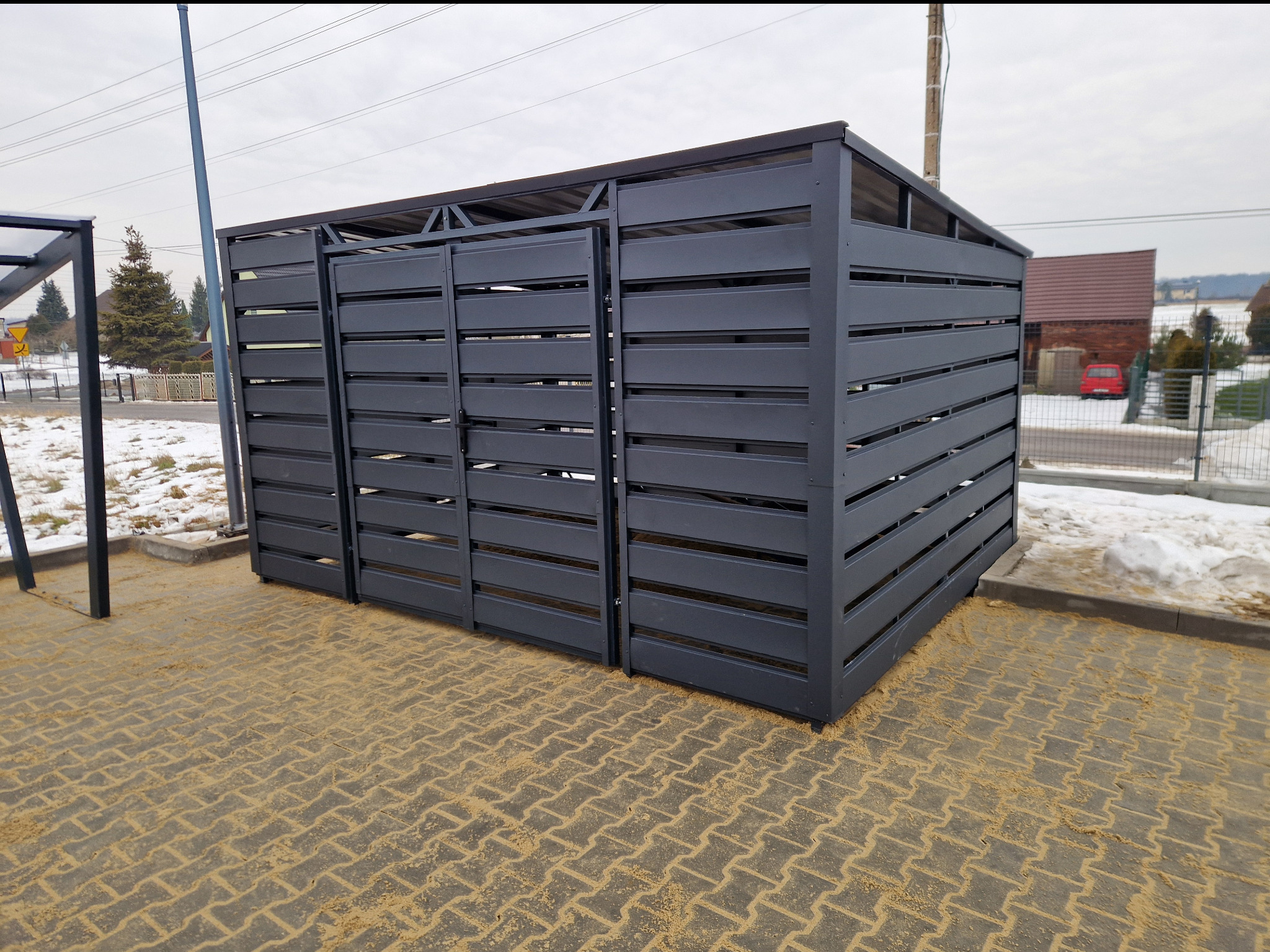 Nowoczesna, metalowa zabudowa śmietnikowa z poziomymi panelami, pomalowana na szaro, na tle kostki brukowej i zimowego krajobrazu.