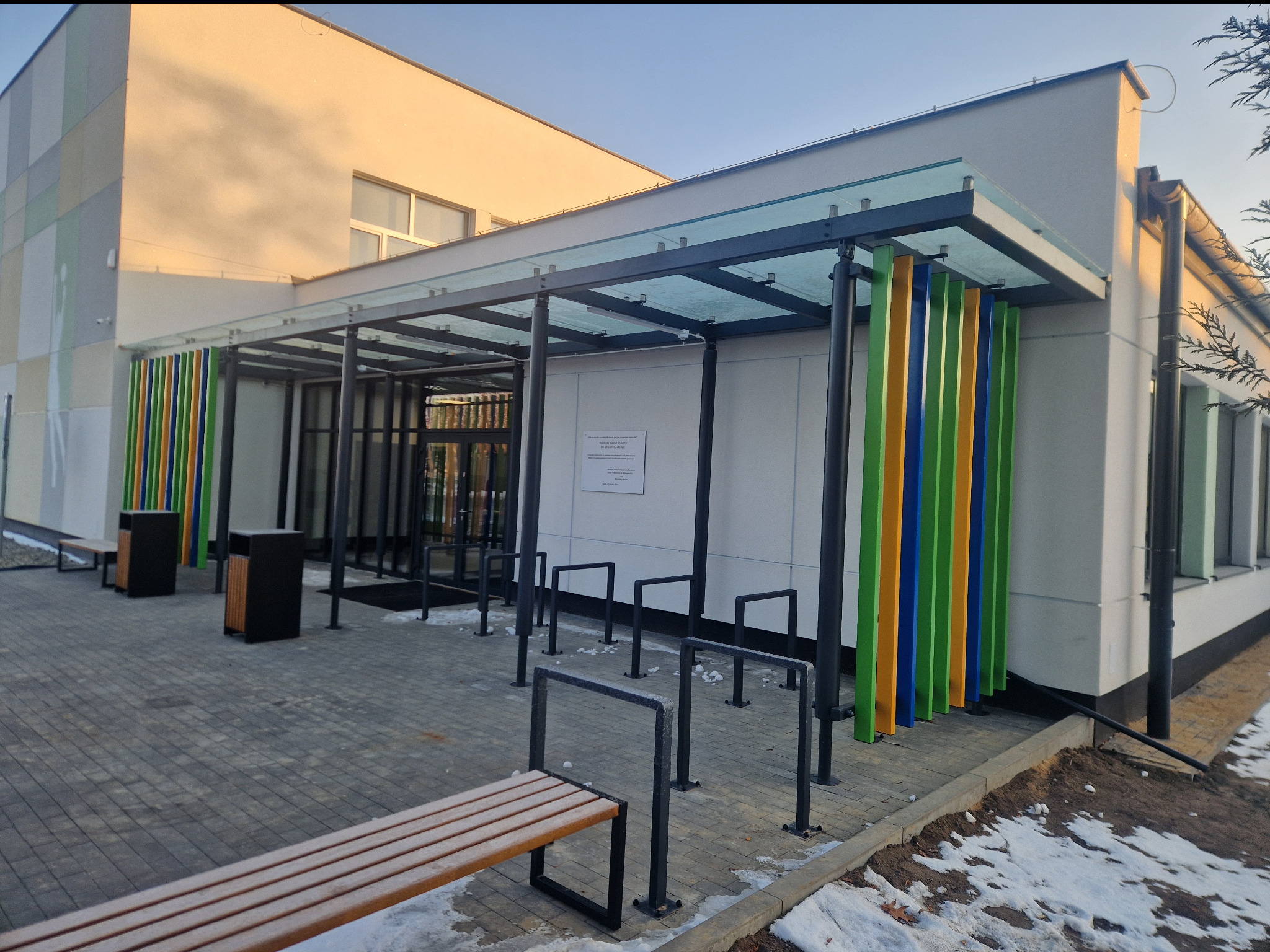 Nowoczesne zadaszenie wejścia do budynku z kolorowymi, pionowymi elementami dekoracyjnymi. Widoczne metalowe stojaki na rowery i ławka.