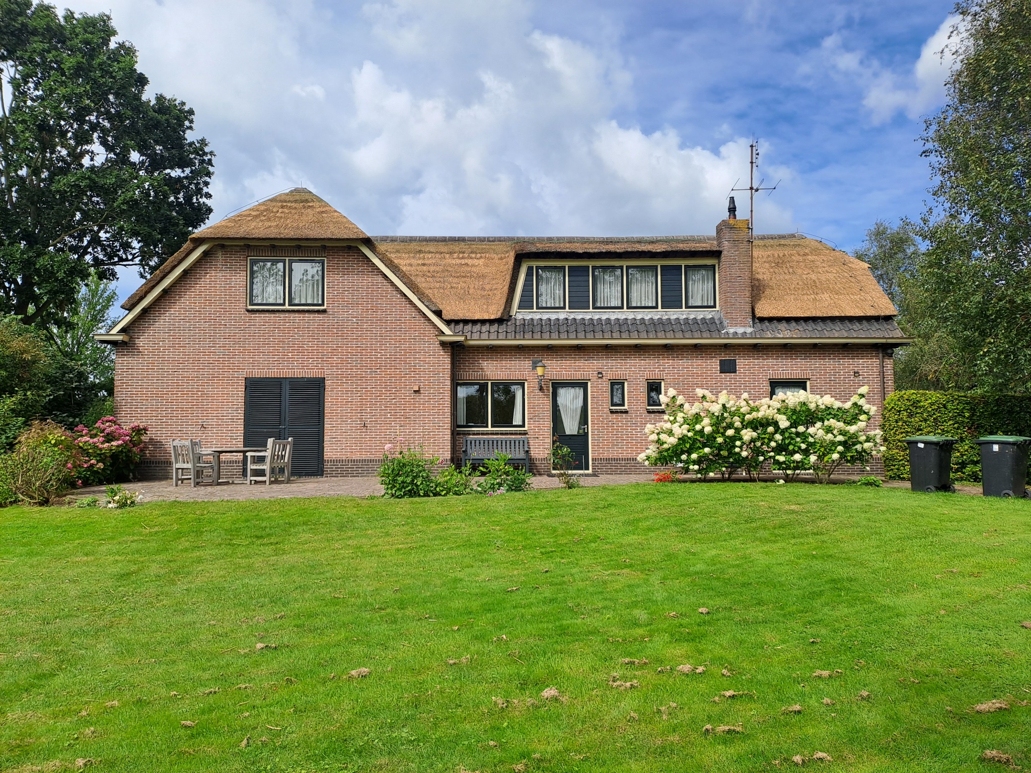 Dom z cegły z krytym strzechą dachem w Trzebnicy, widok z zadbanego trawnika, z przodu białe hortensje i dwa czarne kosze na śmieci.