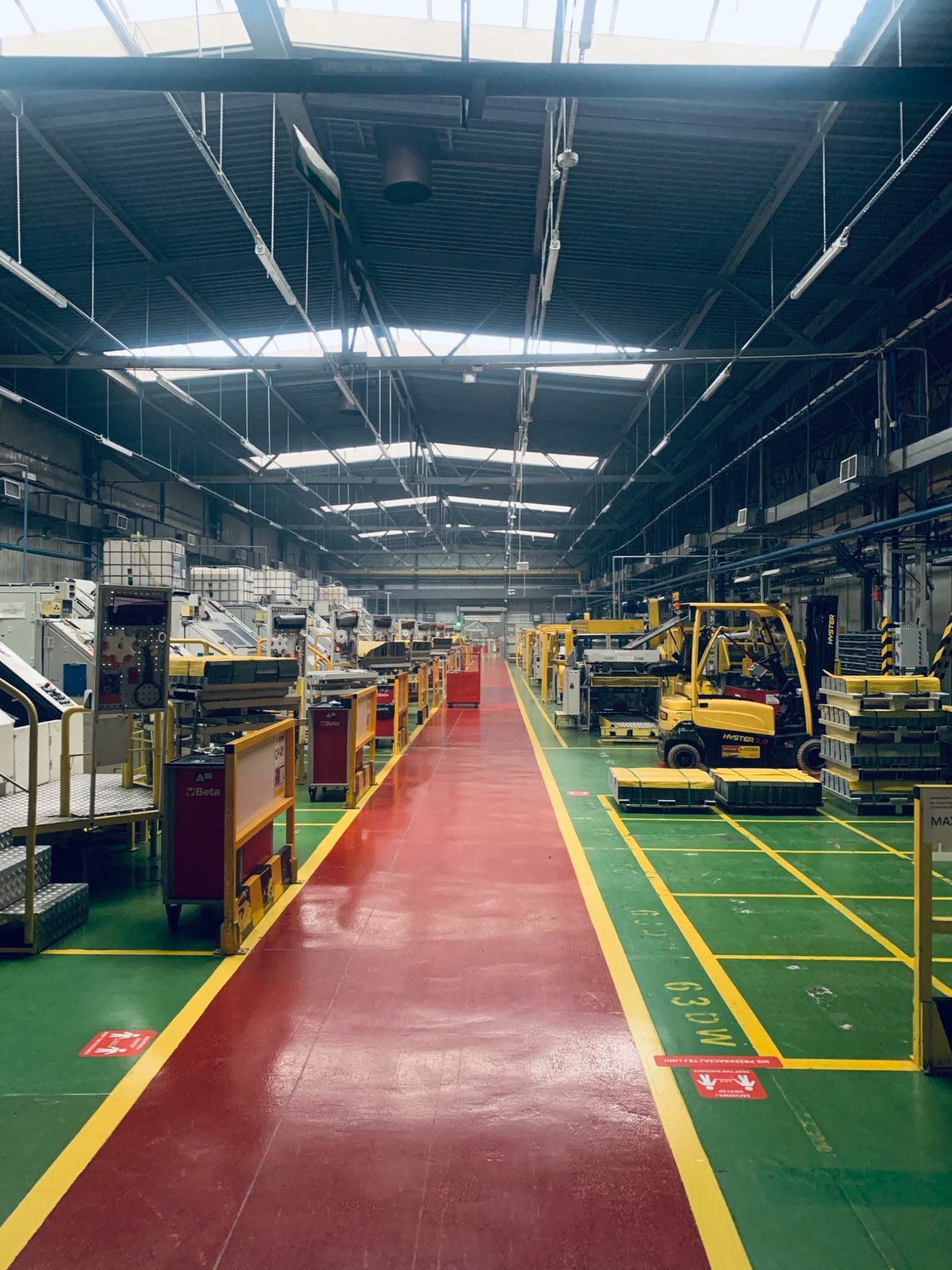 Wnętrze hali produkcyjnej z żółtym wózkiem widłowym Hyster, maszynami i czerwono-zieloną posadzką z wyznaczonymi strefami.