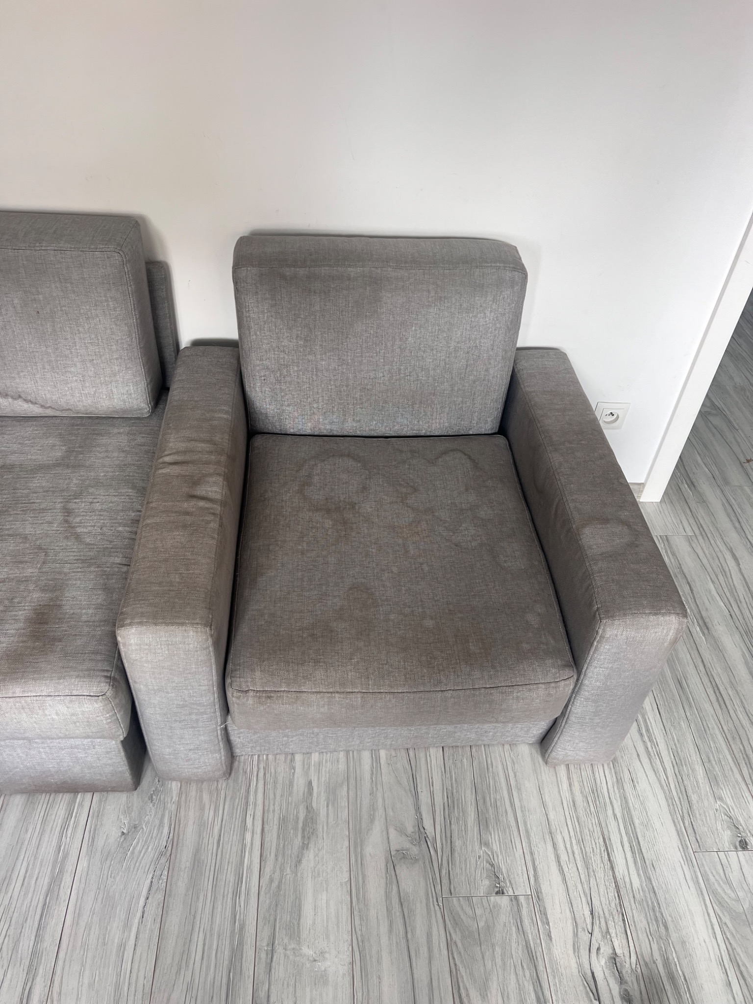 Szary fotel z widocznymi plamami na siedzisku i podłokietnikach, ustawiony na podłodze imitującej drewno, z fragmentem sofy po lewej stronie.