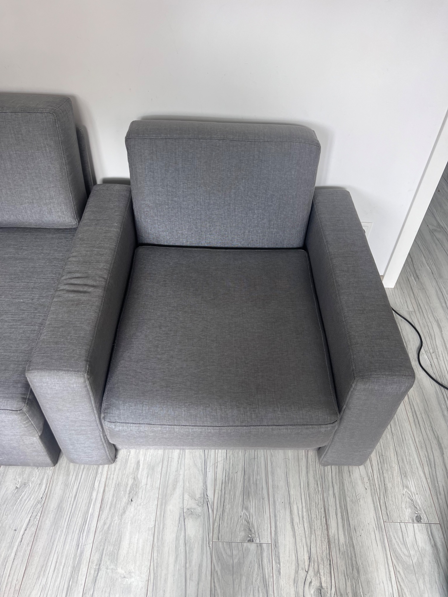 Dwa szare, nowoczesne fotele tapicerowane z widoczną teksturą materiału, ustawione na podłodze imitującej drewno, z białymi ścianami w tle.