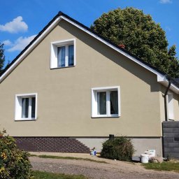 GEBAR HOUSE - kompleksowe wykończenia i remonty - Patryk Gębarowski - Elewacja domu po renowacji: beżowy tynk, białe okna, ciemna podmurówka z dekoracyjnym wzorem. Widoczne puszki po farbie, sugerujące świeżo zakończone prace.