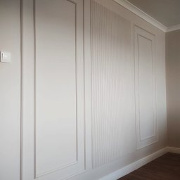 GEBAR HOUSE - kompleksowe wykończenia i remonty - Patryk Gębarowski - Biała ściana z dekoracyjnymi panelami w stylu angielskim, z subtelnym oświetleniem i ciemną podłogą. Wykończenie wnętrza z dbałością o detale.