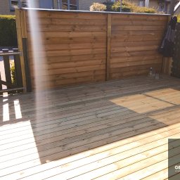 GEBAR HOUSE - kompleksowe wykończenia i remonty - Patryk Gębarowski - Drewniany taras z brązową, poziomą deską ogrodzeniową i czarnym ażurowym ogrodzeniem. Widoczne promienie słońca padające na deski. W tle zielona roślinność.