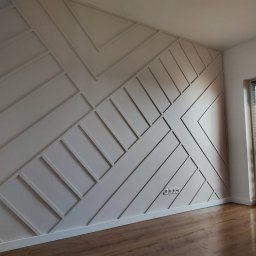 GEBAR HOUSE - kompleksowe wykończenia i remonty - Patryk Gębarowski - Ściana z dekoracyjnymi, geometrycznymi panelami MDF w kolorze jasnego beżu, z widocznymi gniazdkami elektrycznymi i fragmentem podłogi z paneli drewnopodobnych, oświetlona naturalnym światłem...