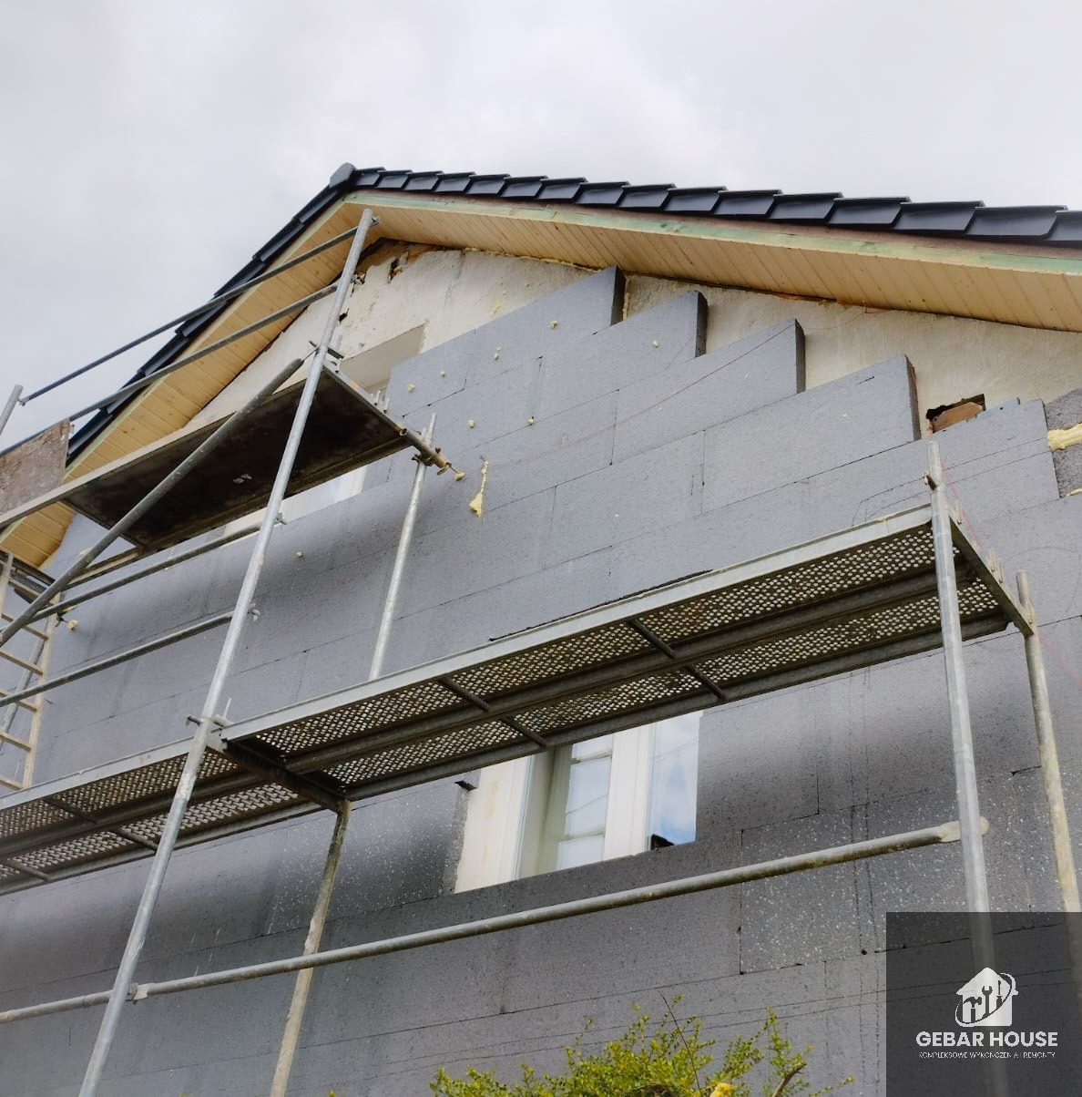 Ocieplanie ściany zewnętrznej domu styropianem grafitowym, widoczne rusztowanie i fragment dachu z czarną dachówką. Etap prac przed nałożeniem tynku.