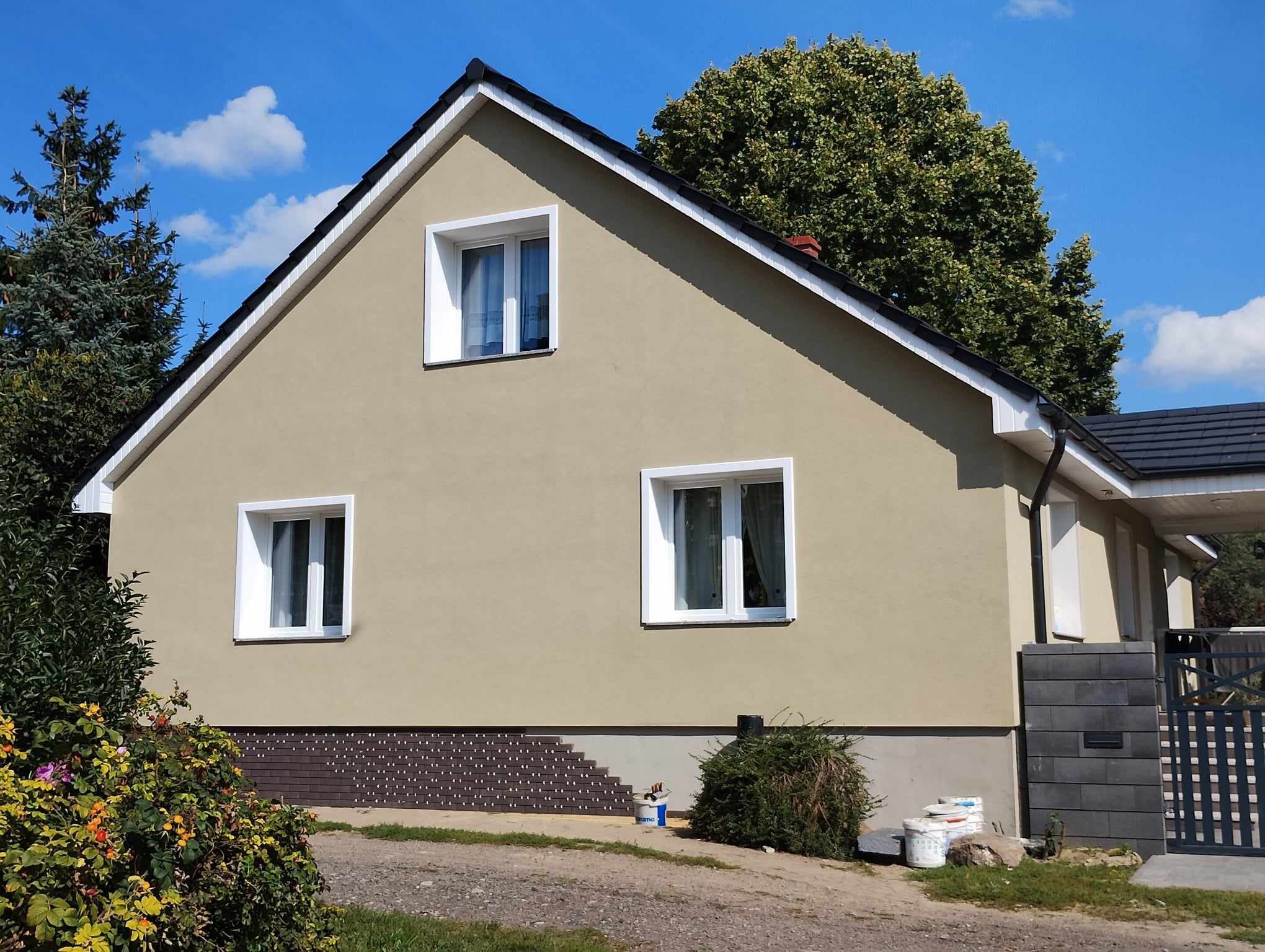 Elewacja domu po renowacji: beżowy tynk, białe okna, ciemna podmurówka z dekoracyjnym wzorem. Widoczne puszki po farbie, sugerujące świeżo zakończone prace.