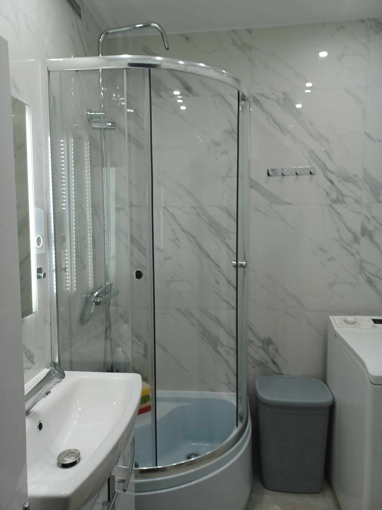 Nowoczesna łazienka po remoncie: kabina prysznicowa z chromowanymi elementami, umywalka z szafką, marmurowe płytki na ścianach, pralka i szary kosz na pranie.