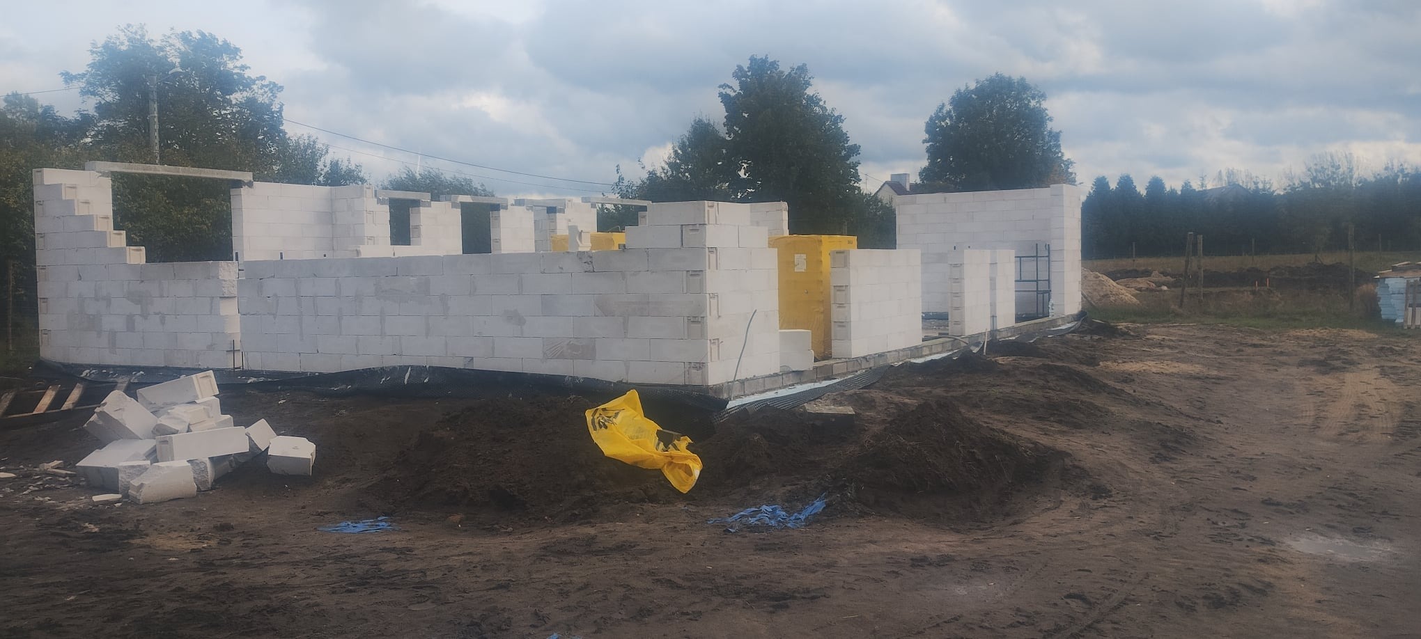 Wznoszenie murów domu z betonu komórkowego, widoczne żółte paczki z materiałem izolacyjnym. Teren budowy z ziemią, w tle zieleń drzew i pochmurne niebo.