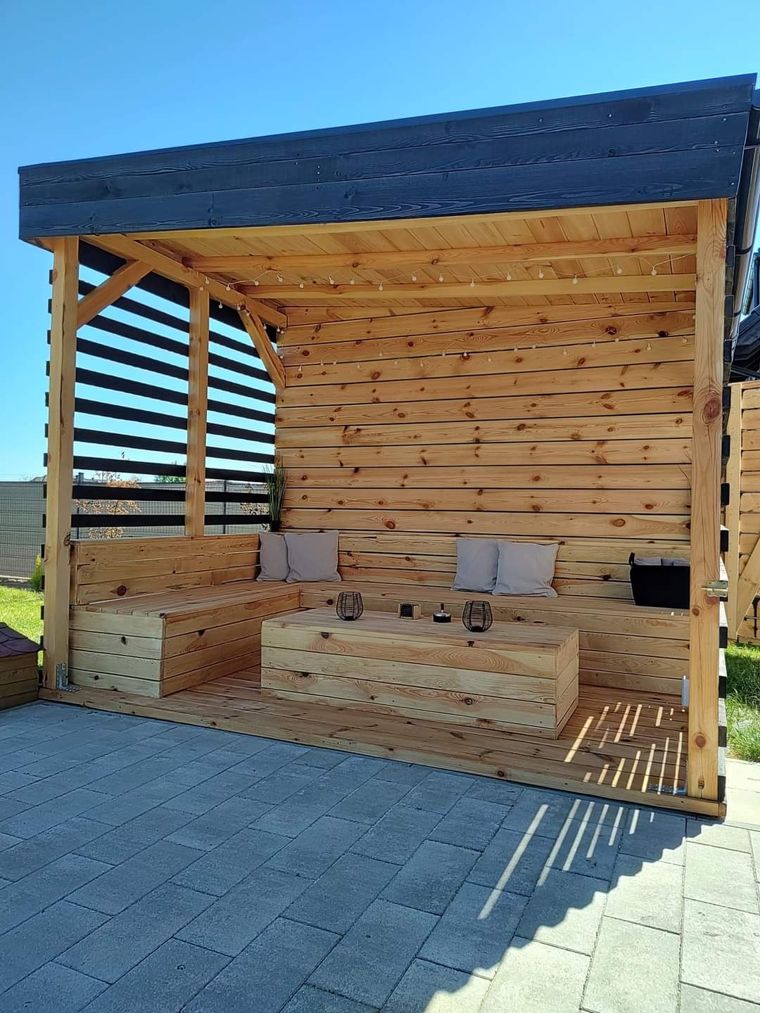 Drewniana altana ogrodowa z meblami, oświetleniem i dekoracjami. Widoczne szare poduszki i elementy dekoracyjne na stole. Całość w jasnych odcieniach drewna.