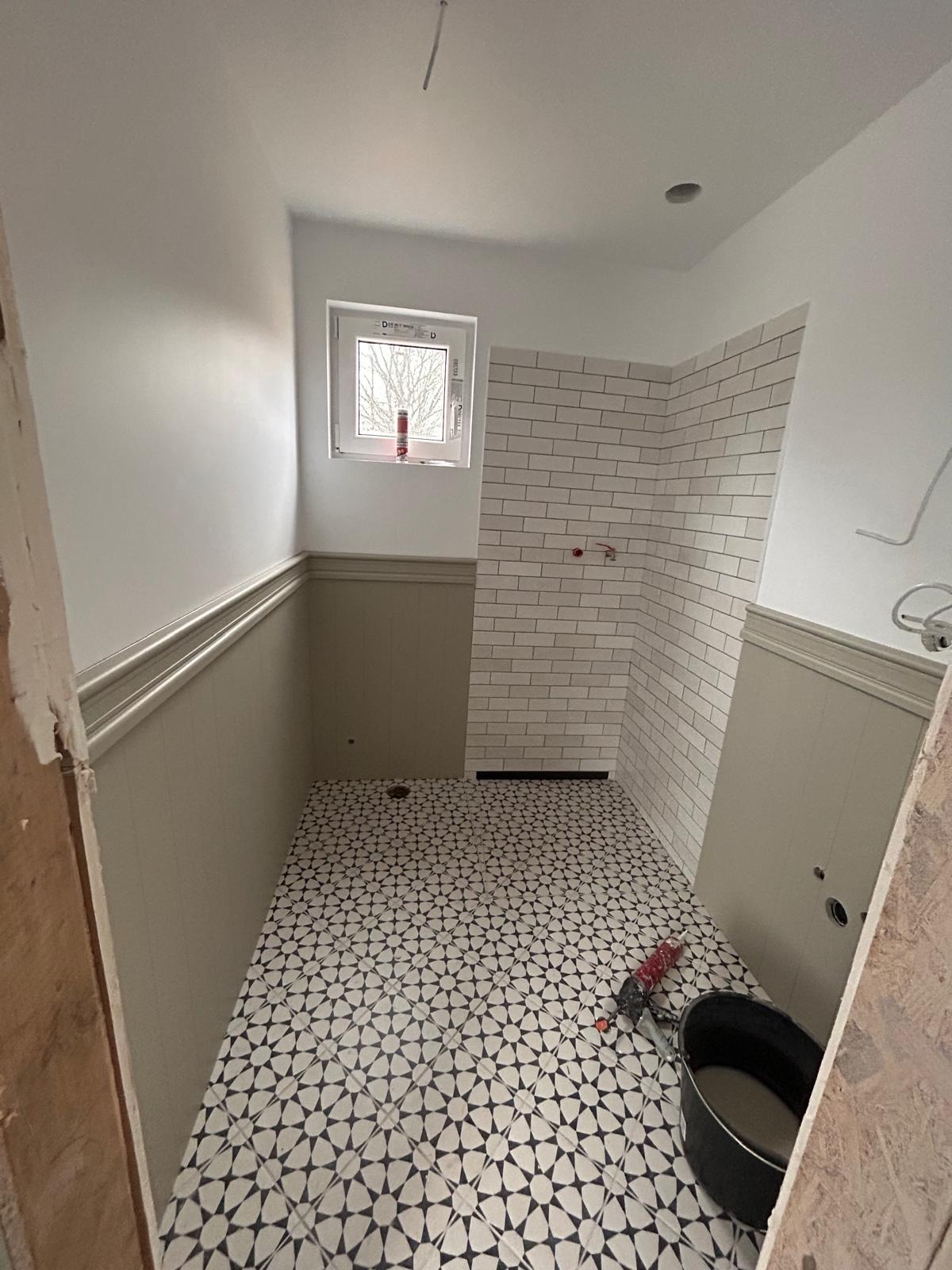 Łazienka w trakcie remontu: białe płytki na ścianach, wzorzysta posadzka, okno z widokiem na drzewa. Widoczne narzędzia i wiadro. Wnętrze w trakcie wykończenia.