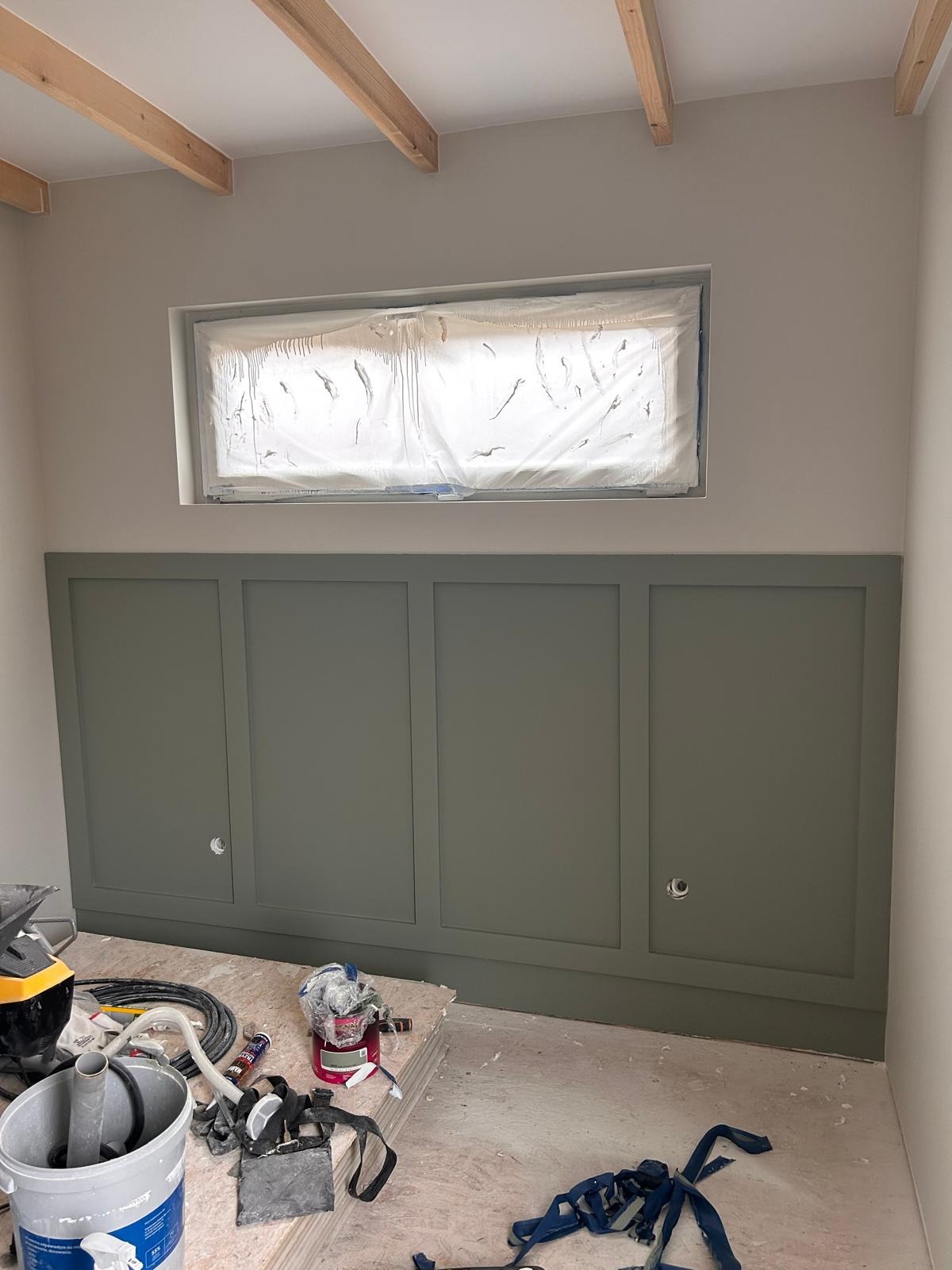 Ściana z panelami w odcieniu zieleni, okno zabezpieczone folią malarską, widoczne narzędzia. Wnętrze w trakcie prac wykończeniowych, belki stropowe na suficie.