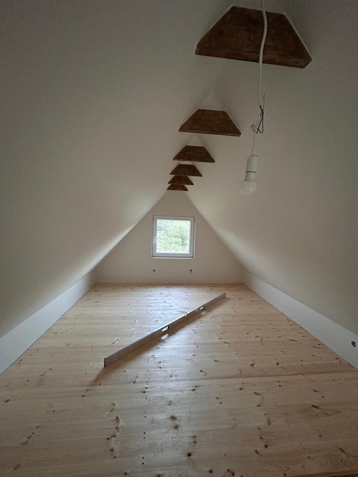 Podłoga z jasnych desek na poddaszu ze skosem, poziomica leży na podłodze, widoczne drewniane elementy dekoracyjne na suficie i okno.