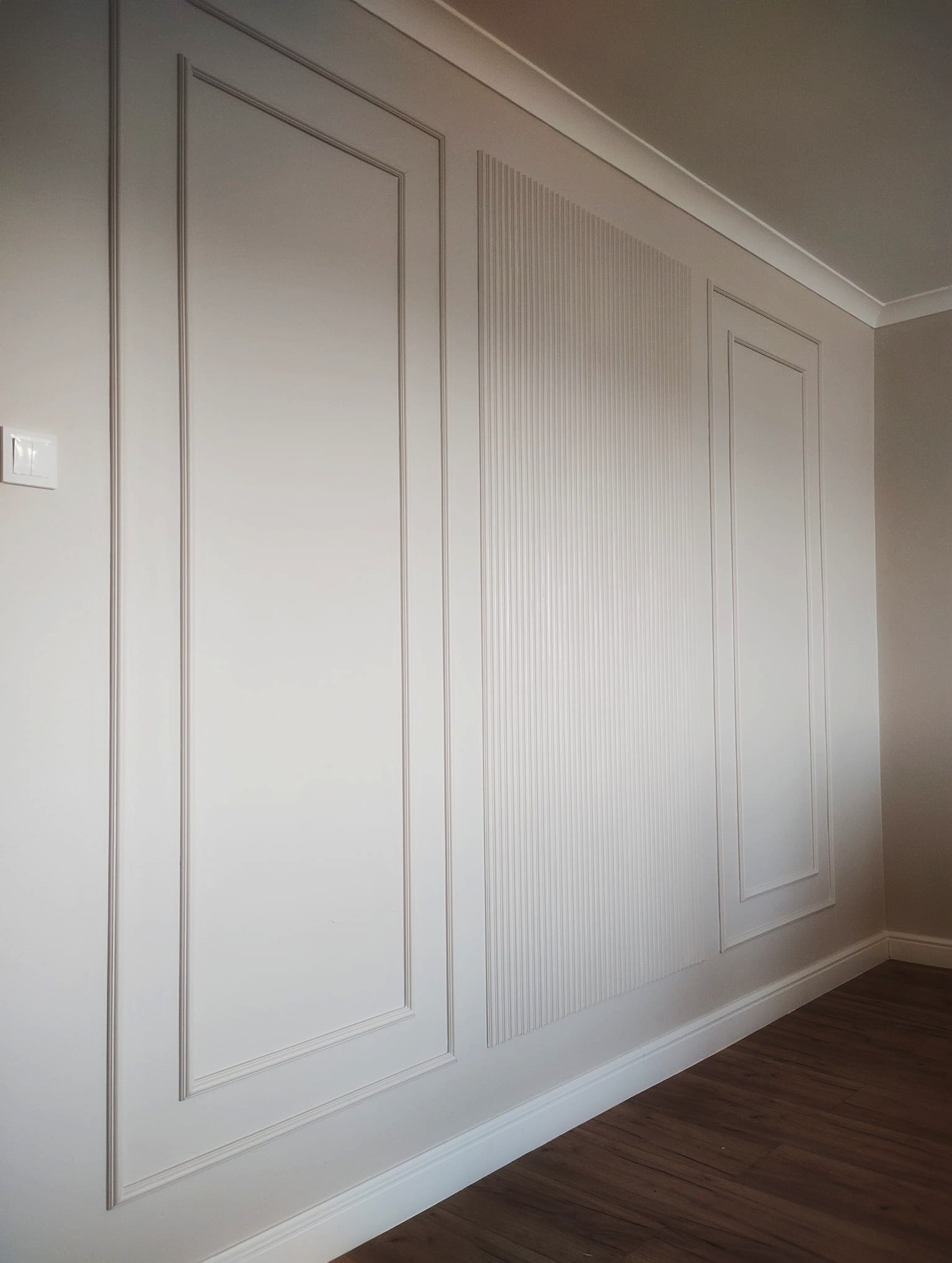 Biała ściana z dekoracyjnymi panelami w stylu angielskim, z subtelnym oświetleniem i ciemną podłogą. Wykończenie wnętrza z dbałością o detale.