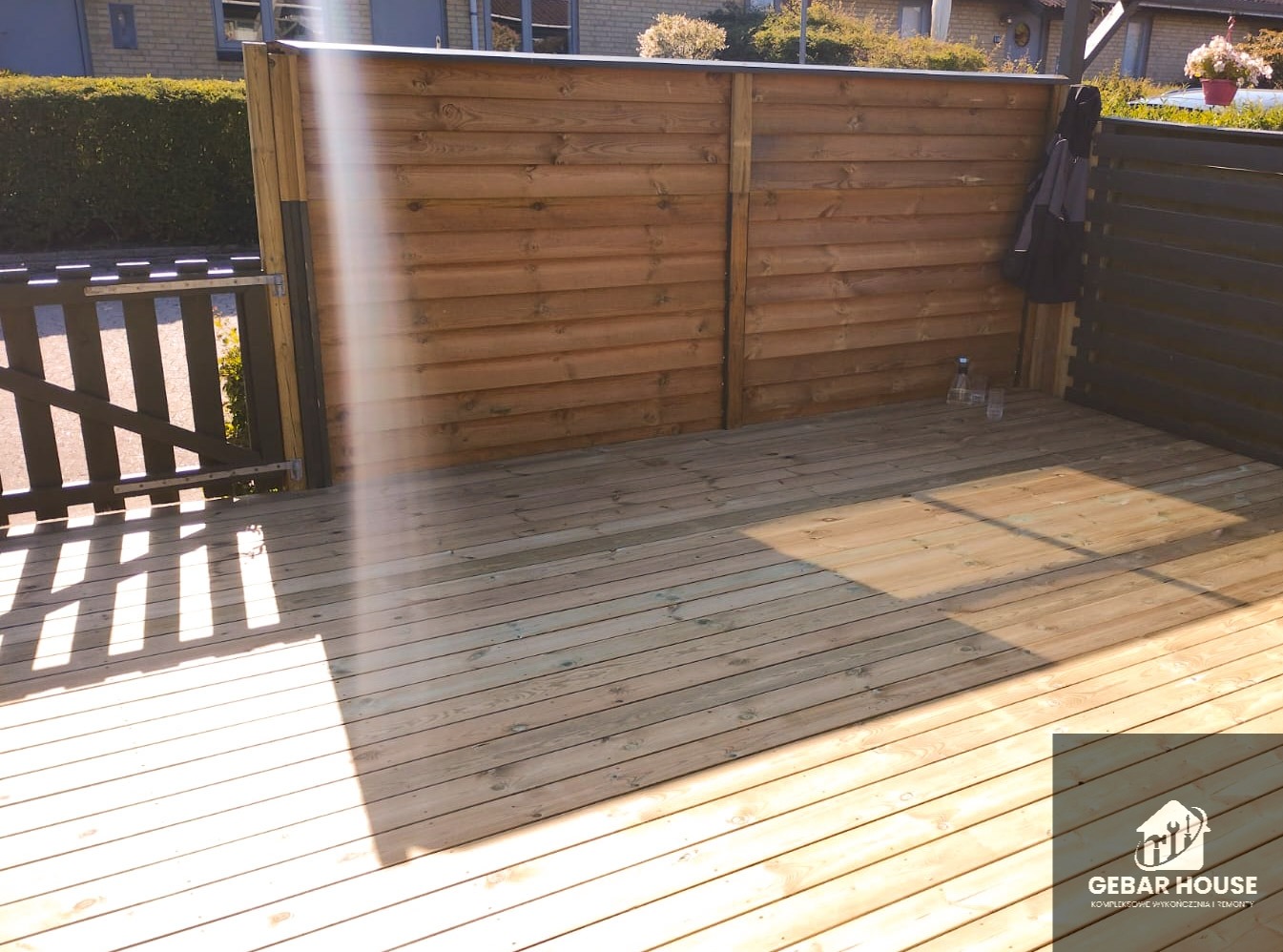 Drewniany taras z brązową, poziomą deską ogrodzeniową i czarnym ażurowym ogrodzeniem. Widoczne promienie słońca padające na deski. W tle zielona roślinność.