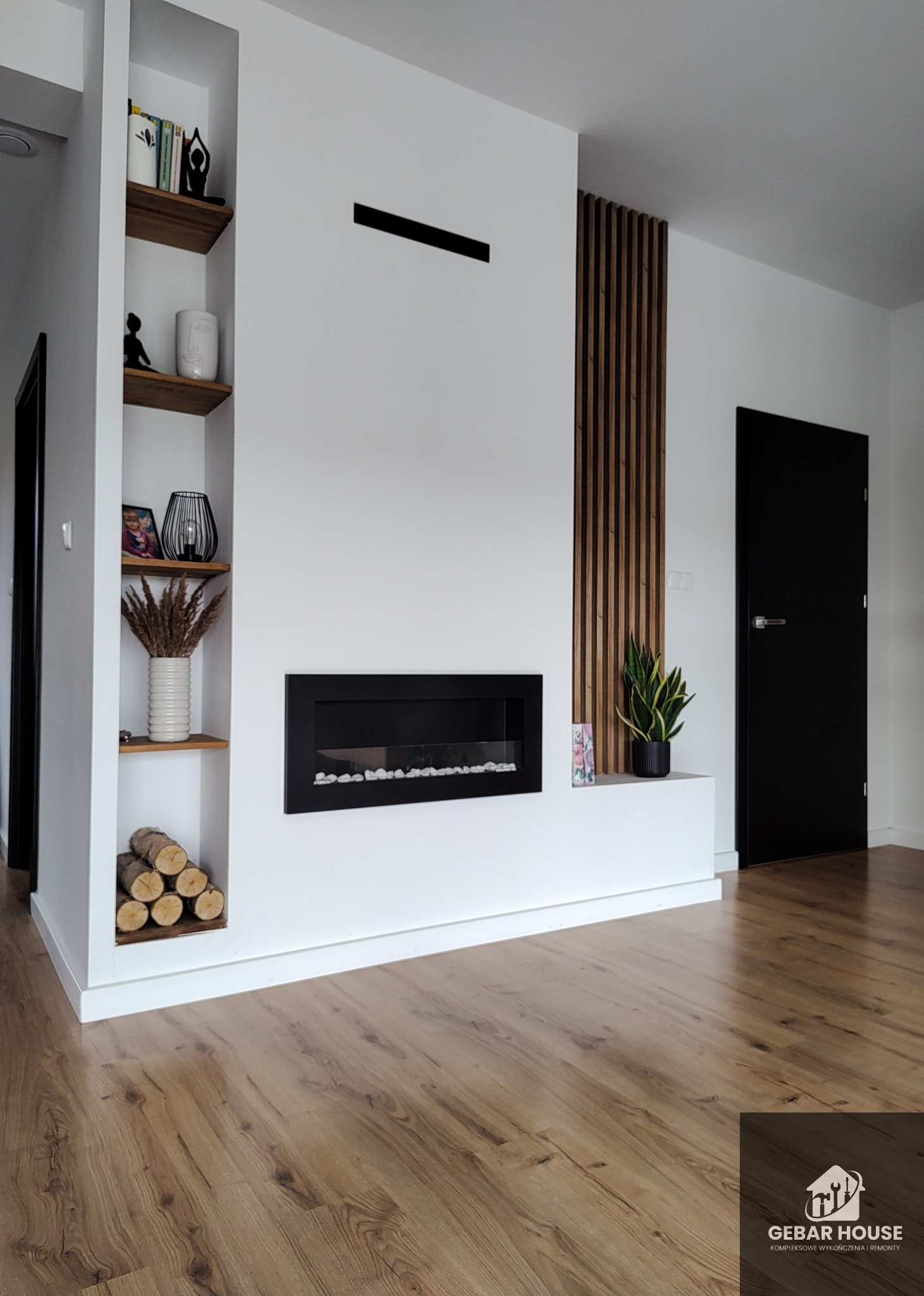 Nowoczesny salon z białymi ścianami, drewnianą podłogą, wbudowanym kominkiem, drewnianymi lamelami dekoracyjnymi i półkami w ścianie z dekoracjami.