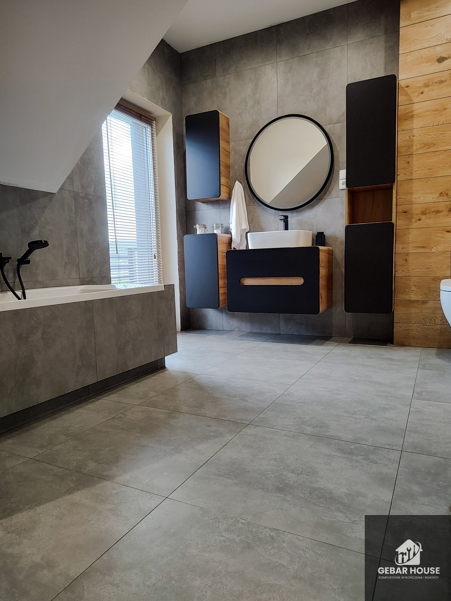 Nowoczesna łazienka w odcieniach szarości z drewnianymi akcentami, okrągłe lustro nad umywalką, wiszące szafki i duża wanna z czarną armaturą, widoczne logo firmy w prawym dolnym rogu.
