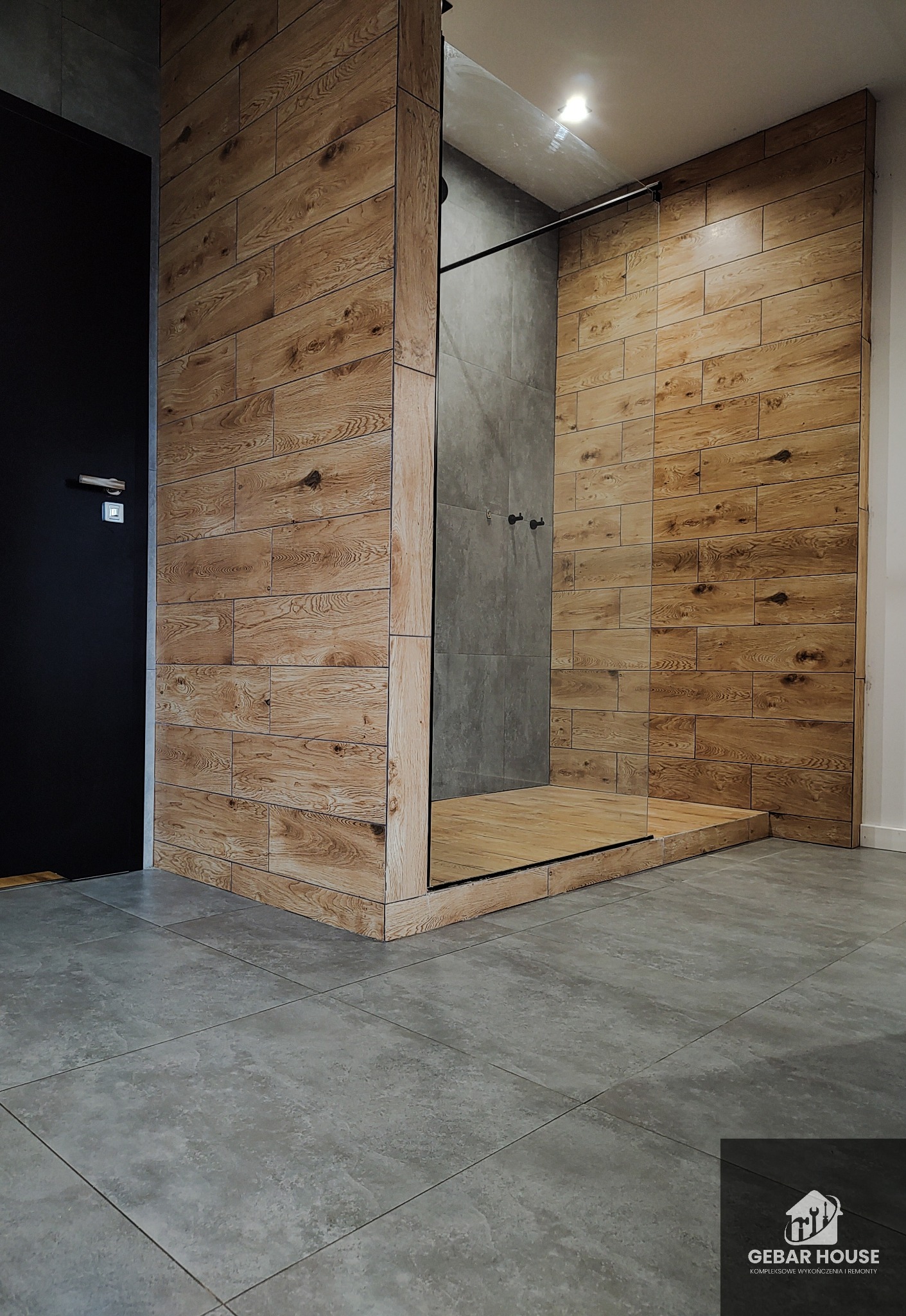 Nowoczesna łazienka z kabiną prysznicową typu walk-in, wykończoną płytkami imitującymi drewno, szara podłoga z dużych płytek, czarne drzwi po lewej stronie.