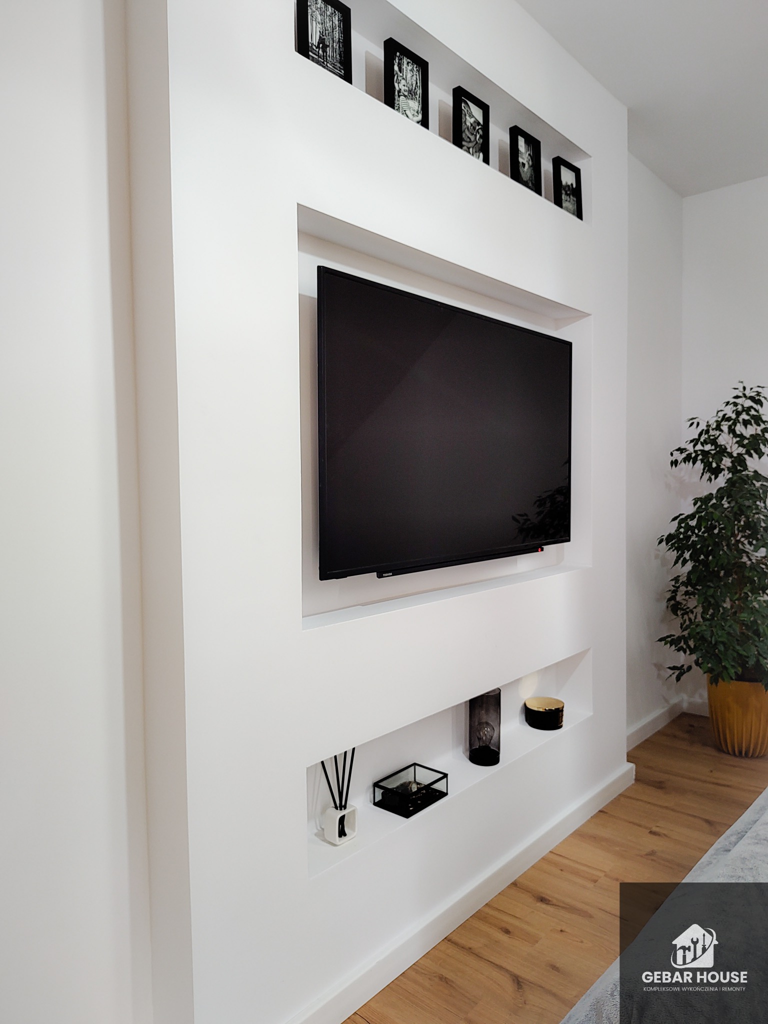 Nowoczesna ściana telewizyjna z wnękami, wykończona gładzią, z czarnymi ramkami zdjęć nad telewizorem i dekoracjami w dolnych wnękach, na podłodze parkiet.