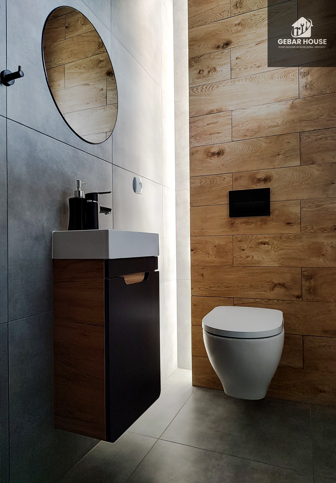 Nowoczesna łazienka z drewnianą ścianą, wiszącą toaletą i umywalką z szafką, okrągłe lustro i szare płytki.